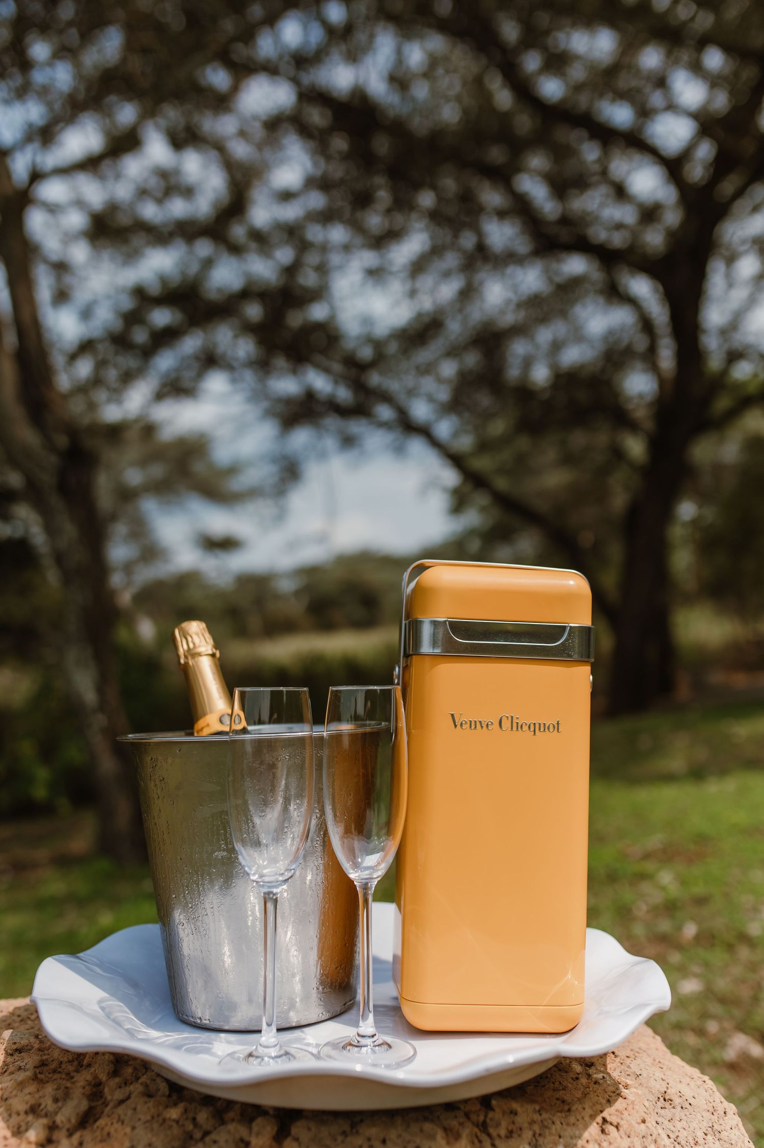 Elegant outdoor champagne setup with Veuve Clicquot box, ice bucket, and crystal glasses on a rustic surface.