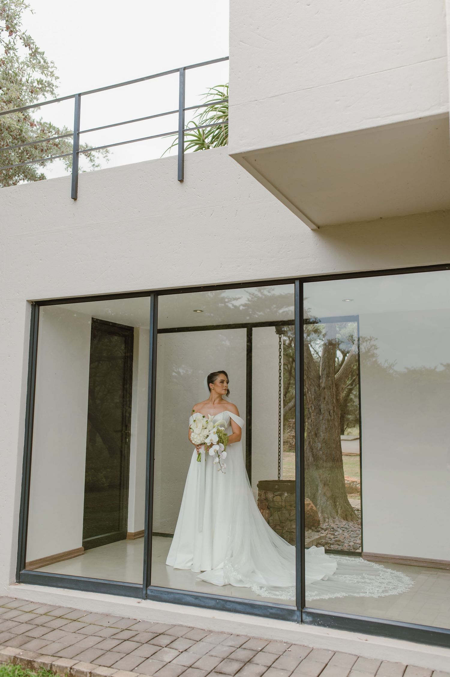 Bride in elegant white wedding gown standing in modern glass-walled room with architectural details.