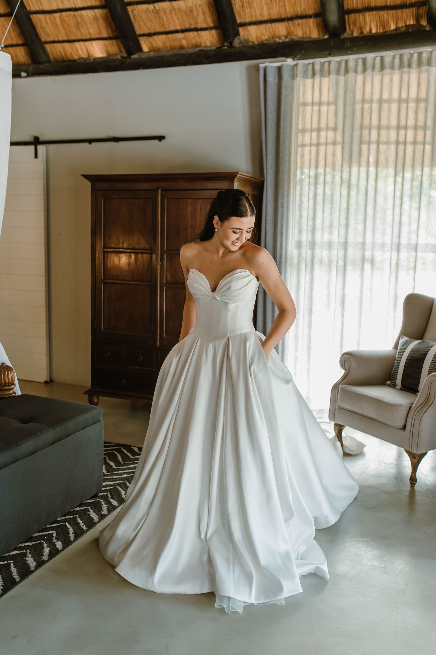 Bride in a strapless sweetheart ballgown wedding dress smiling in a rustic thatched-roof room.