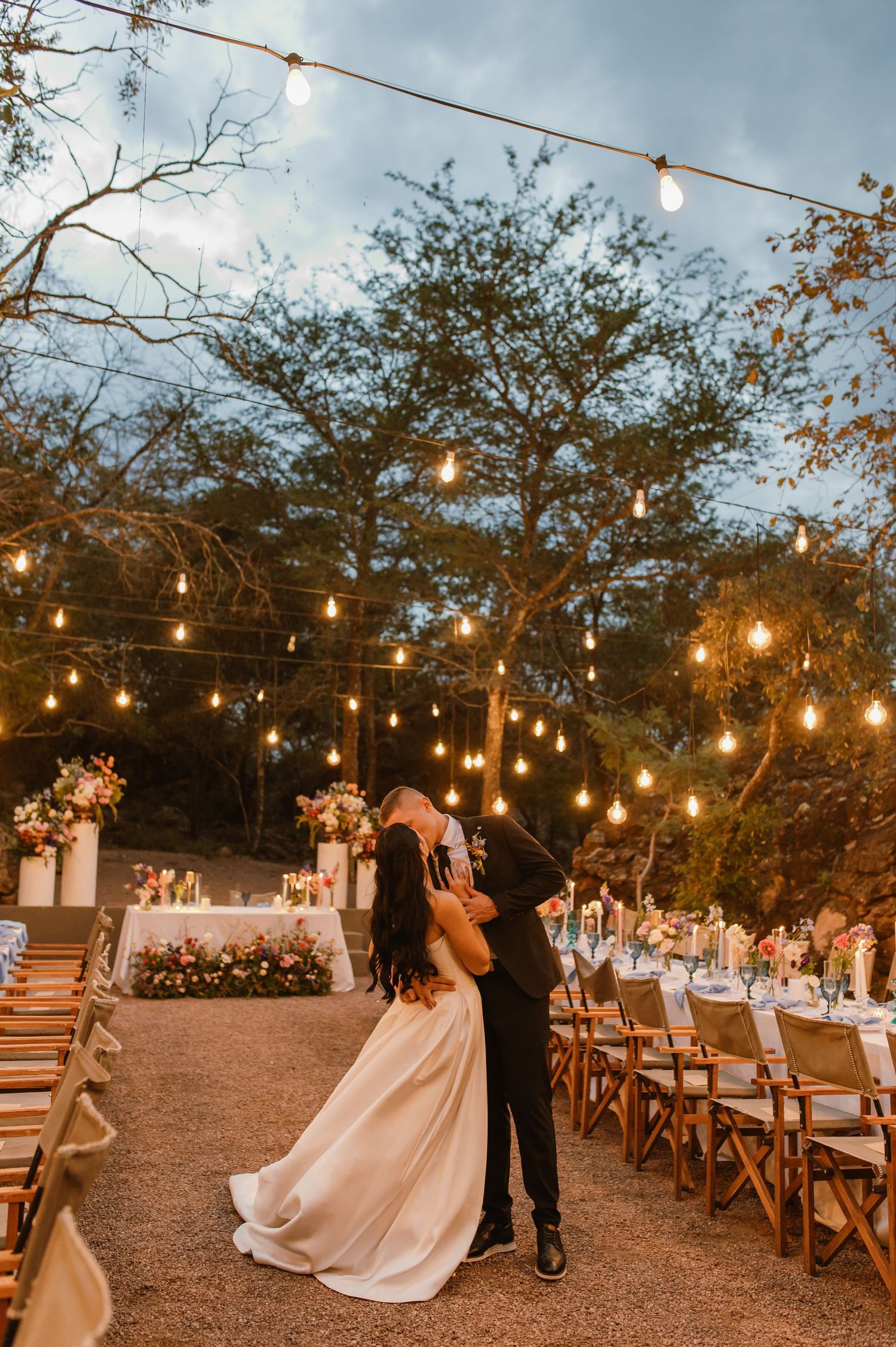 Bride and groom share first dance kiss under glowing string lights at elegant outdoor wedding reception.