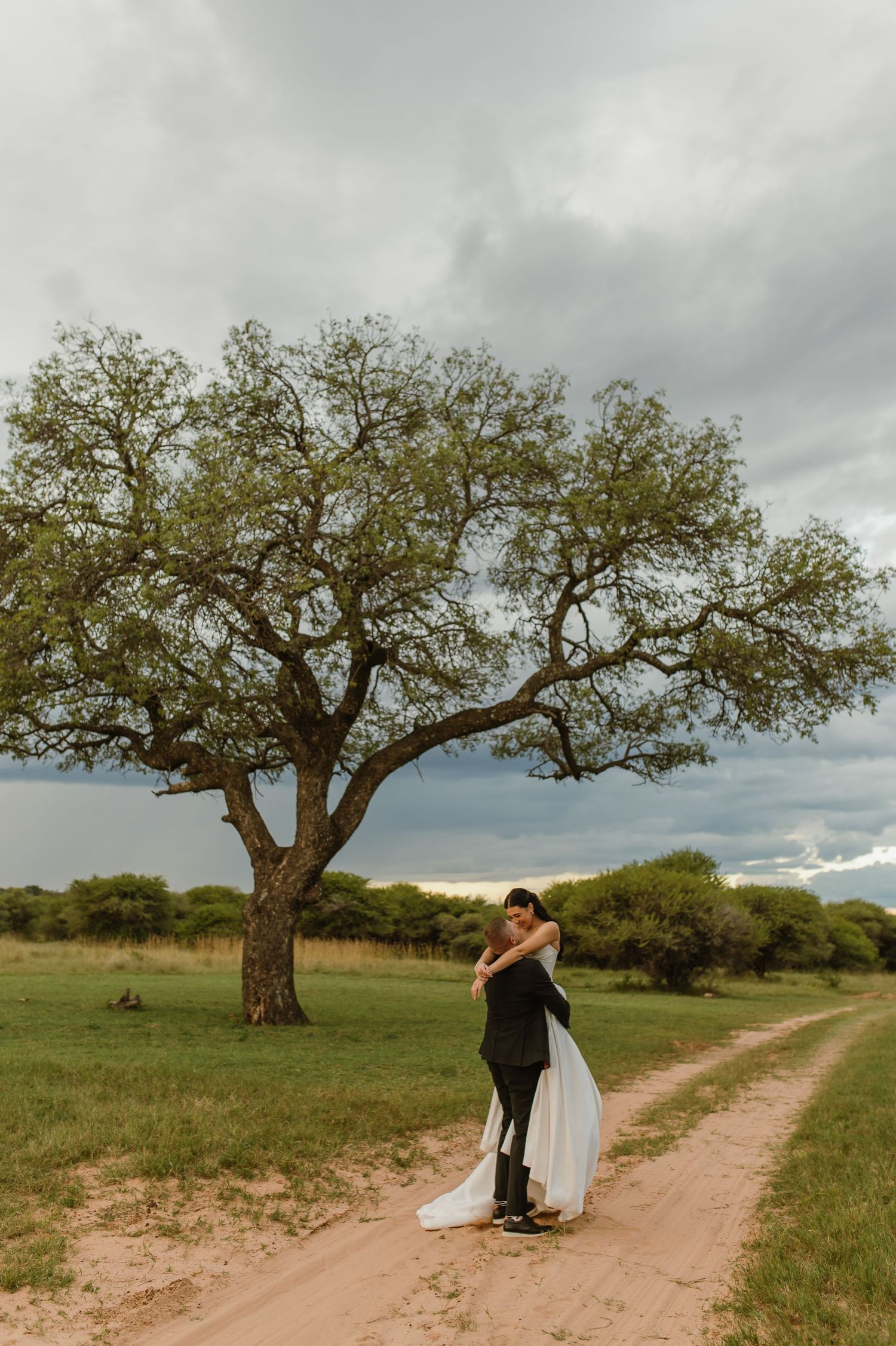 Bride and groom embrace on a dirt path beneath a sprawling tree under dramatic storm clouds.