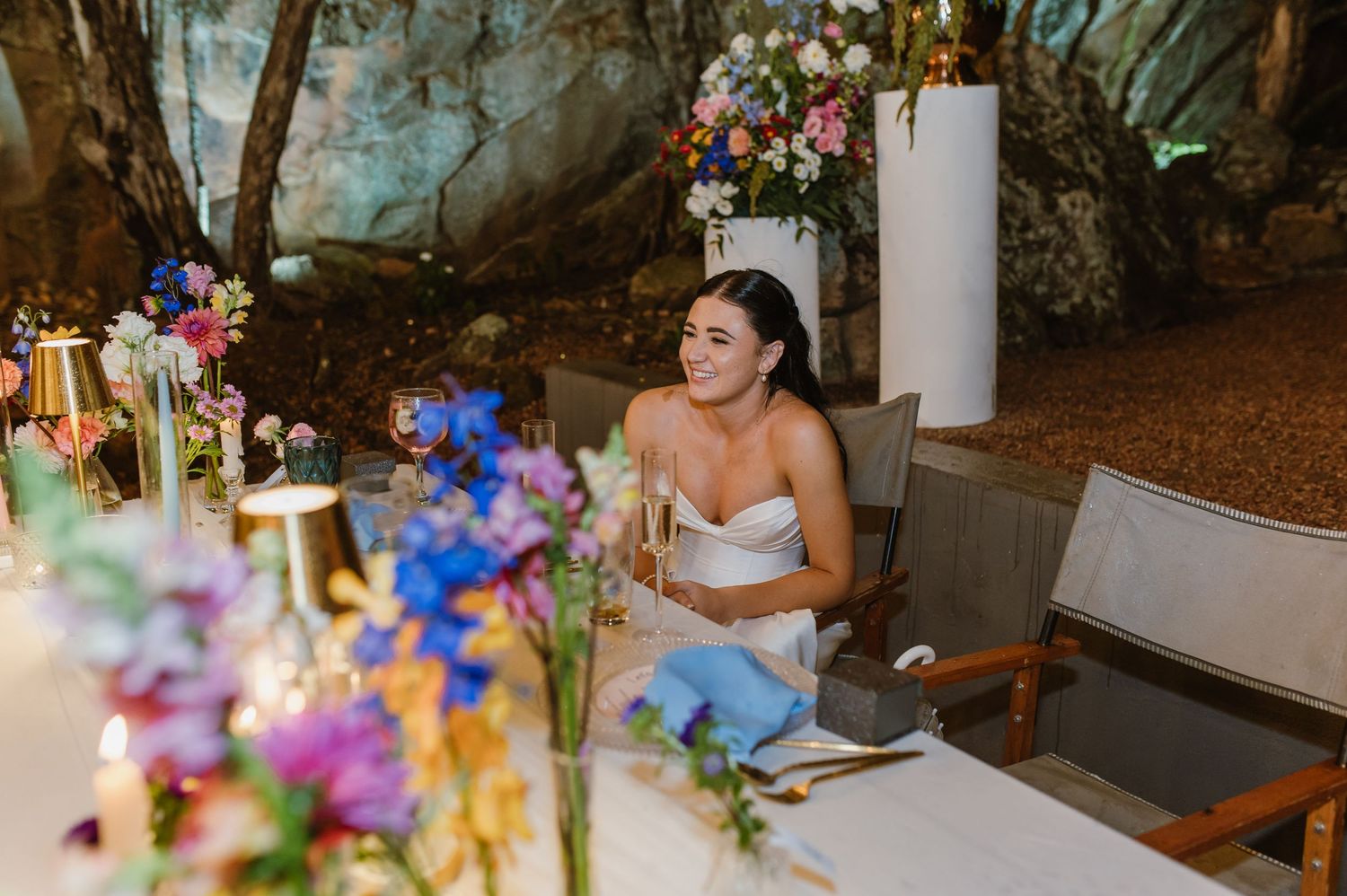 Bride in white gown smiling while holding champagne glass at colorful floral reception table in cave setting.
