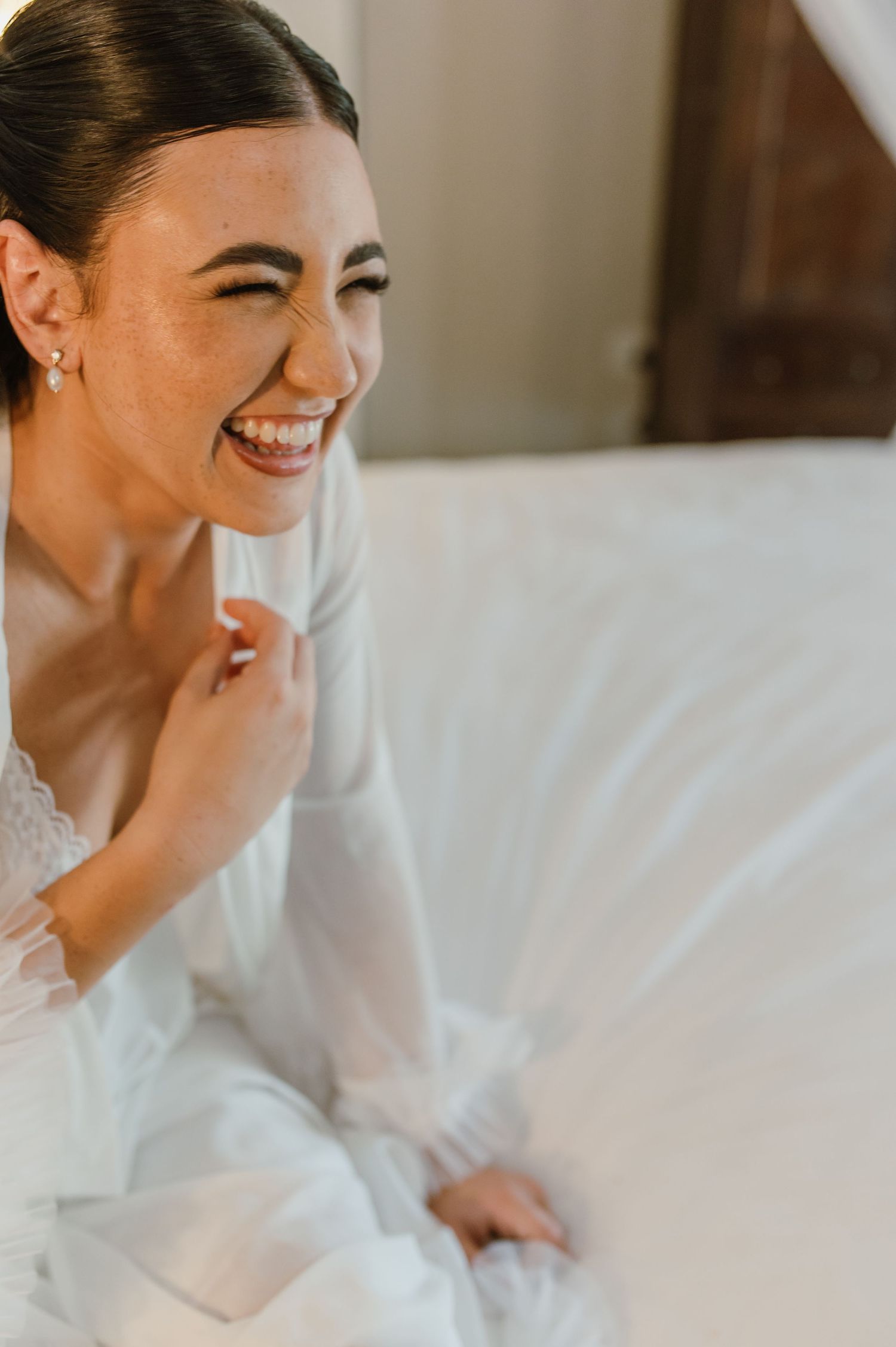 Radiant bride in white lace robe laughing joyfully while getting ready on her wedding day.