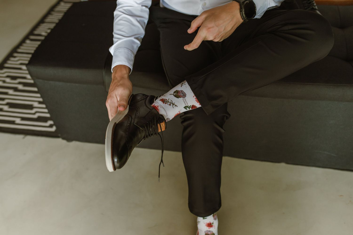 Groom sitting on a couch putting on black dress shoes with fun patterned wedding socks.