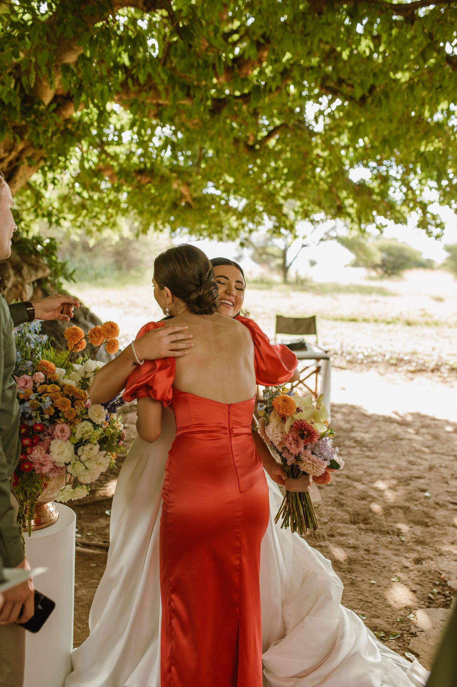 Bride embracing bridesmaid in red dress while holding colorful bouquet at outdoor ceremony.