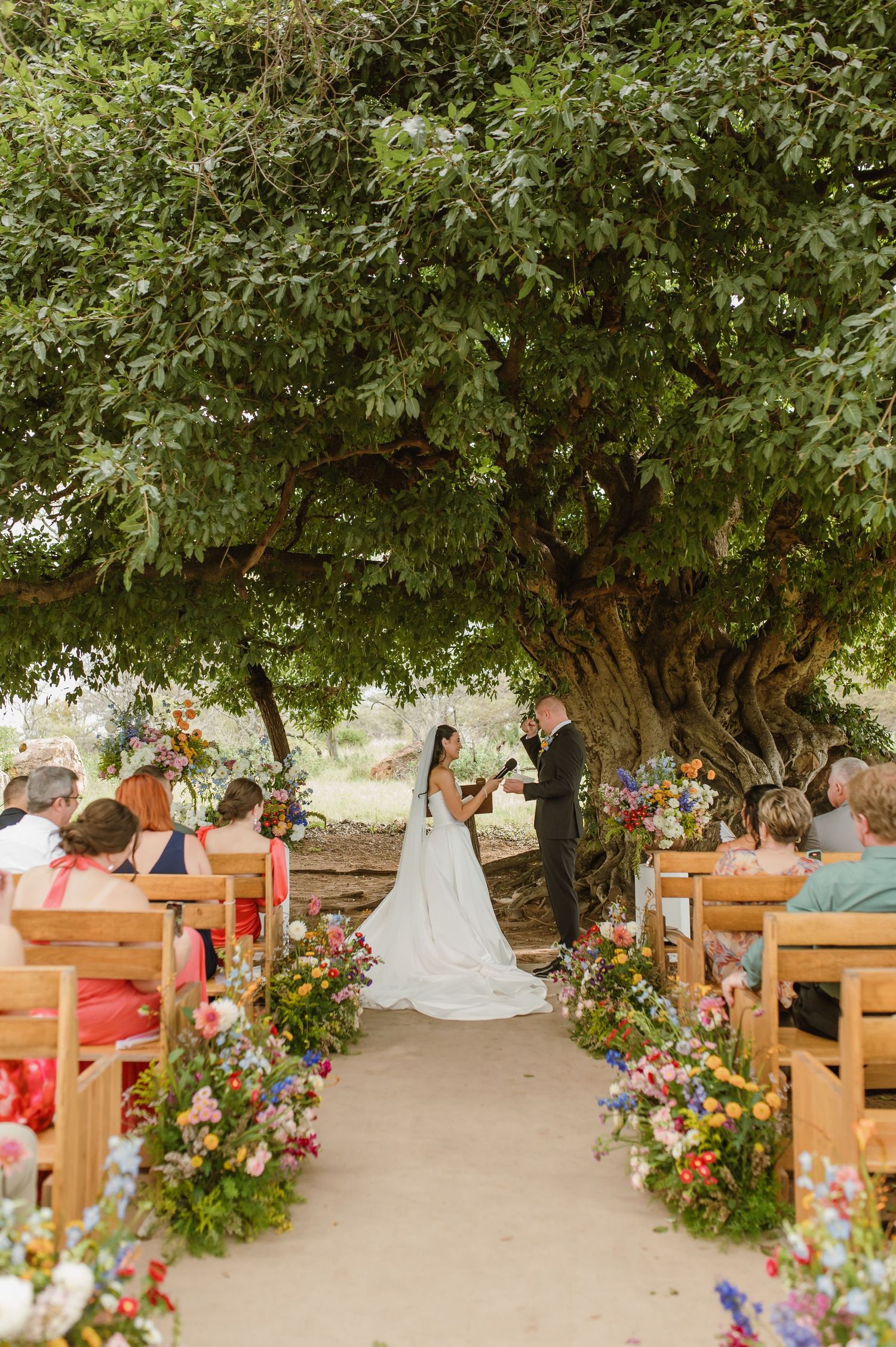 Outdoor wedding ceremony under a massive tree with colorful wildflower arrangements lining the aisle and seated guests.