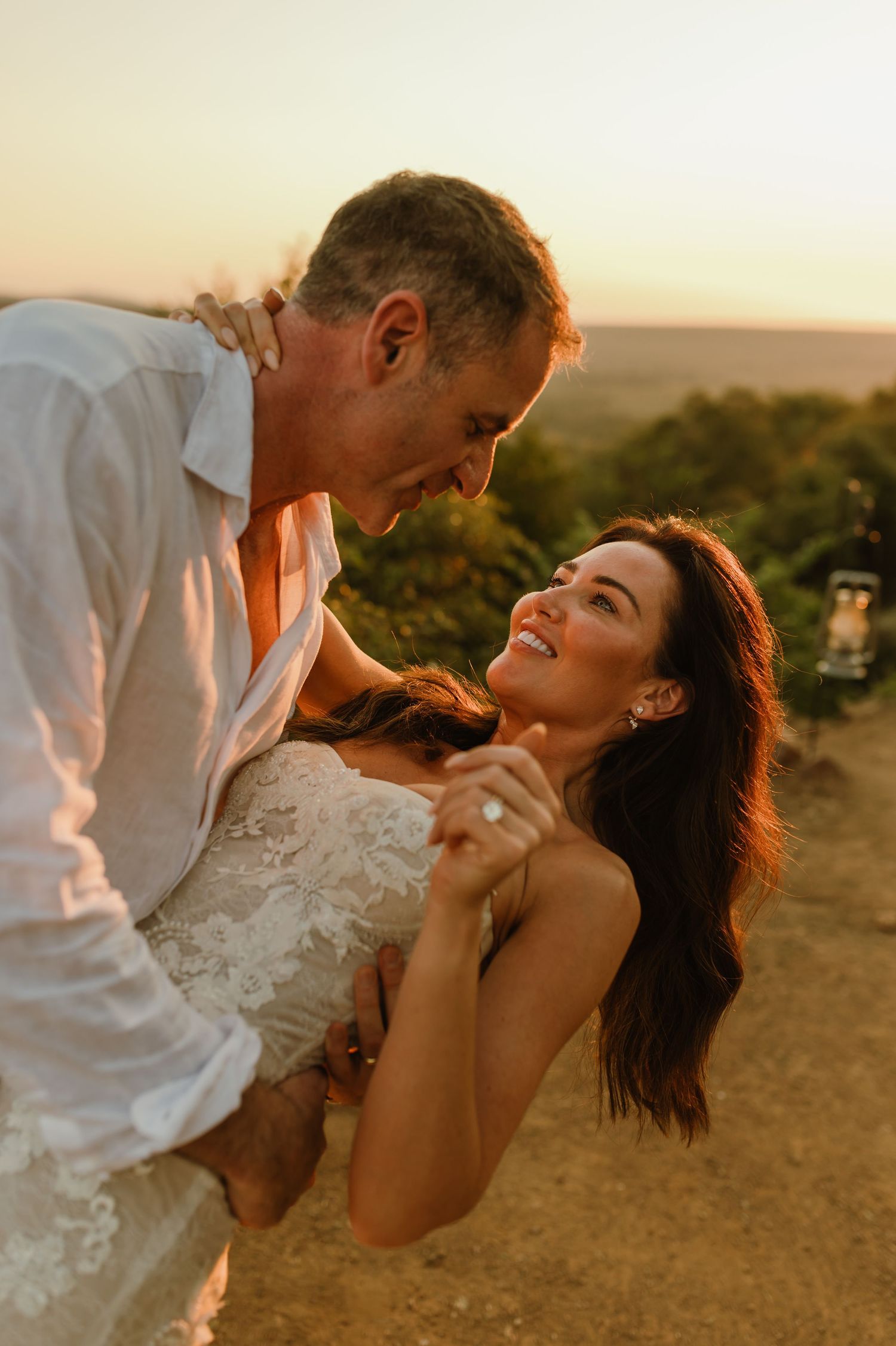 A couple shares a romantic dance at sunset, the bride smiling up at the groom in a lace dress outdoors.