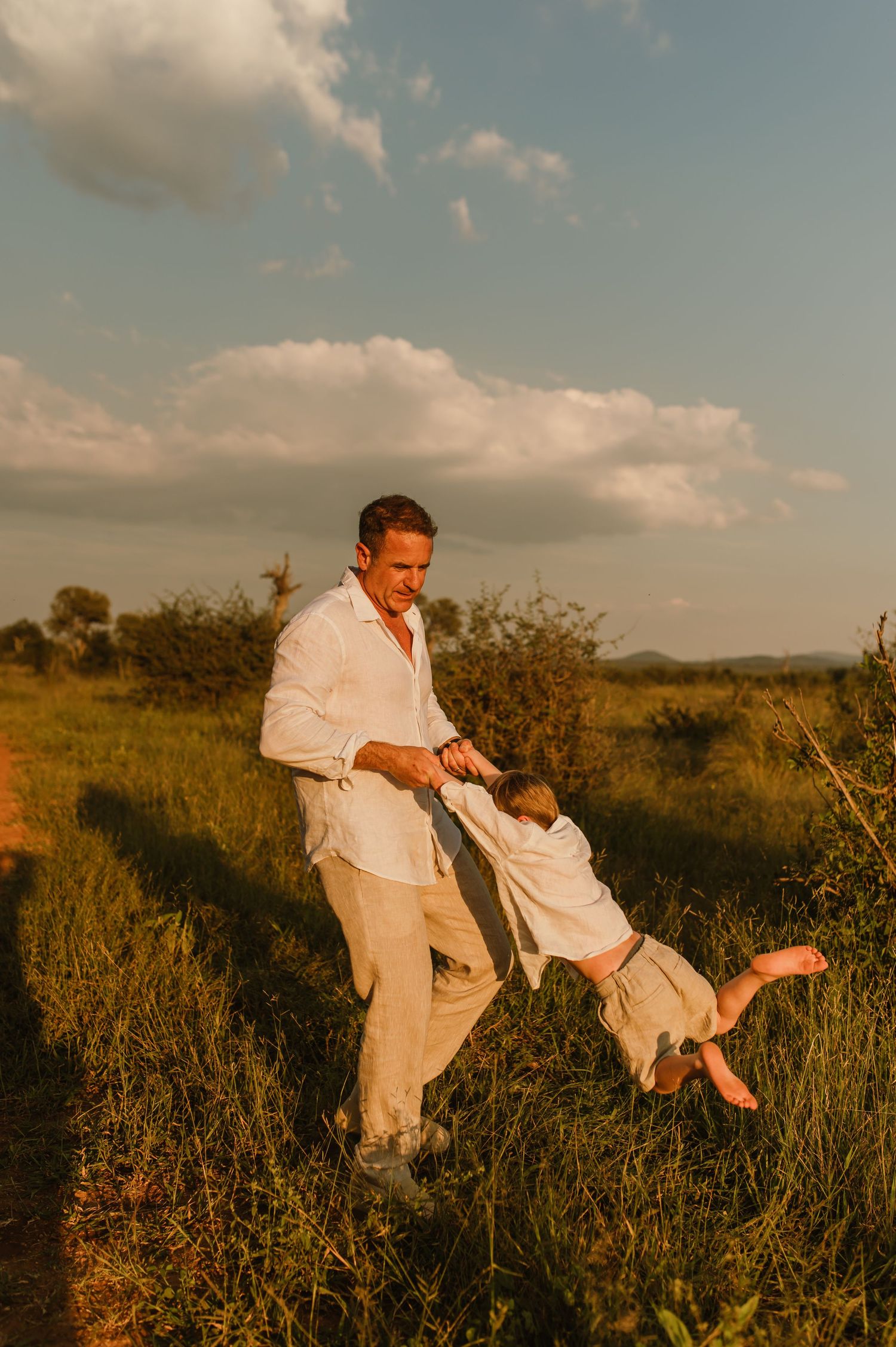 Father playfully swings barefoot child in golden sunset light in a lush green field.
