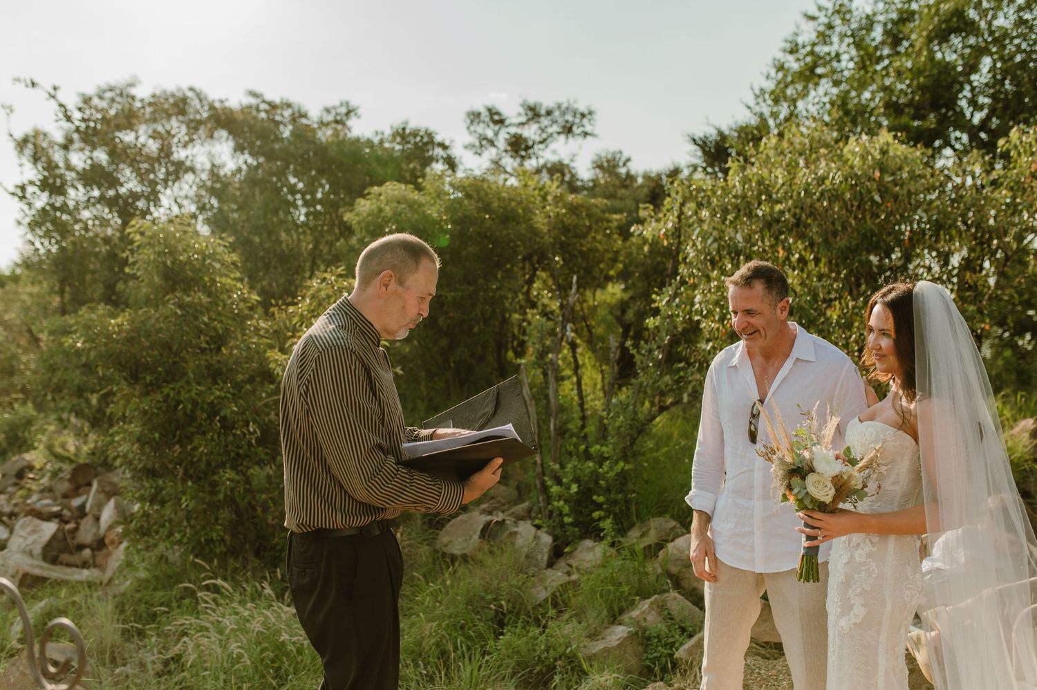 Outdoor wedding ceremony with officiant reading vows to couple in natural garden setting at sunset.
