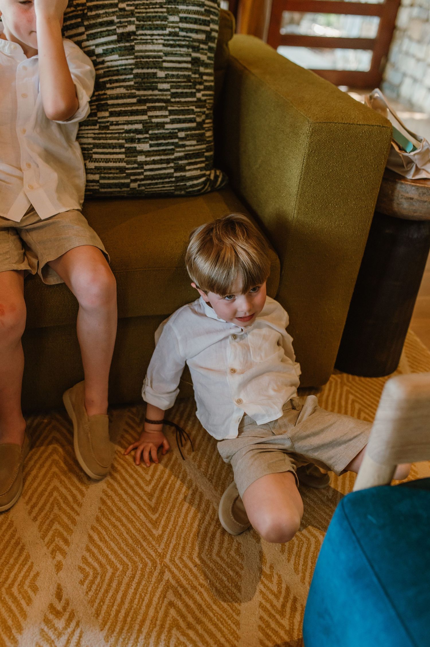 A young boy in a white shirt and tan shorts sits on a patterned carpet beside a green sofa during a wedding.