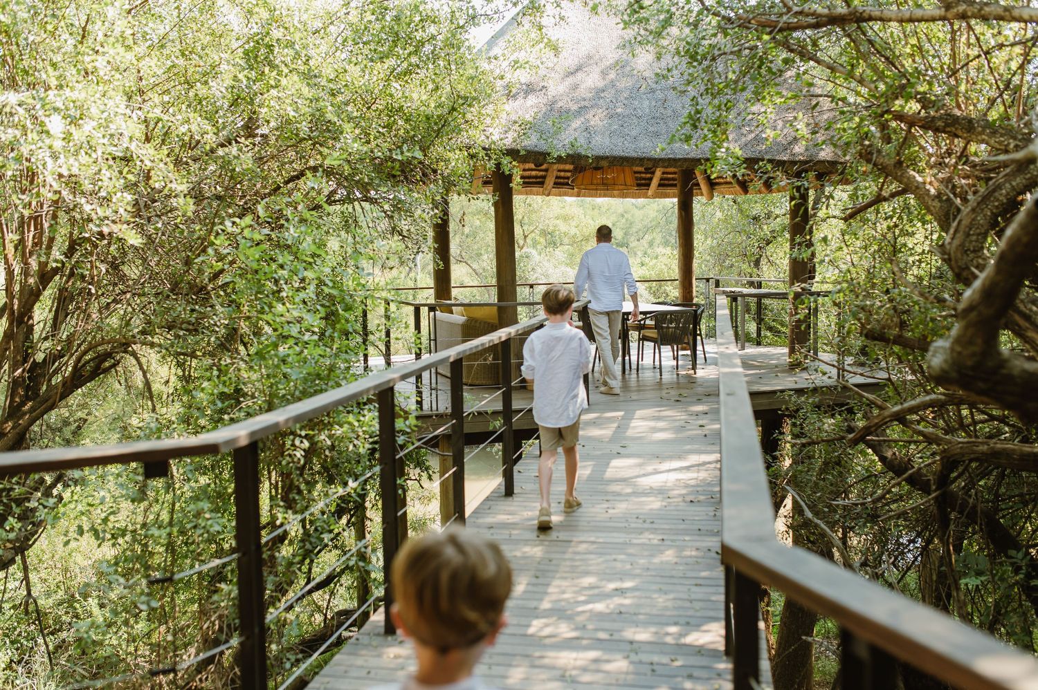Adults and child walking along a wooden boardwalk through lush green trees at a safari lodge.