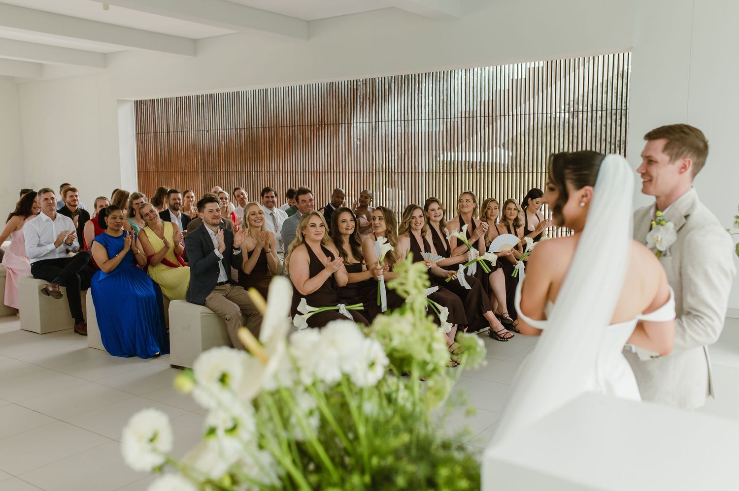 Wedding ceremony in modern white room with blue-dressed guests and floral decorations in foreground.