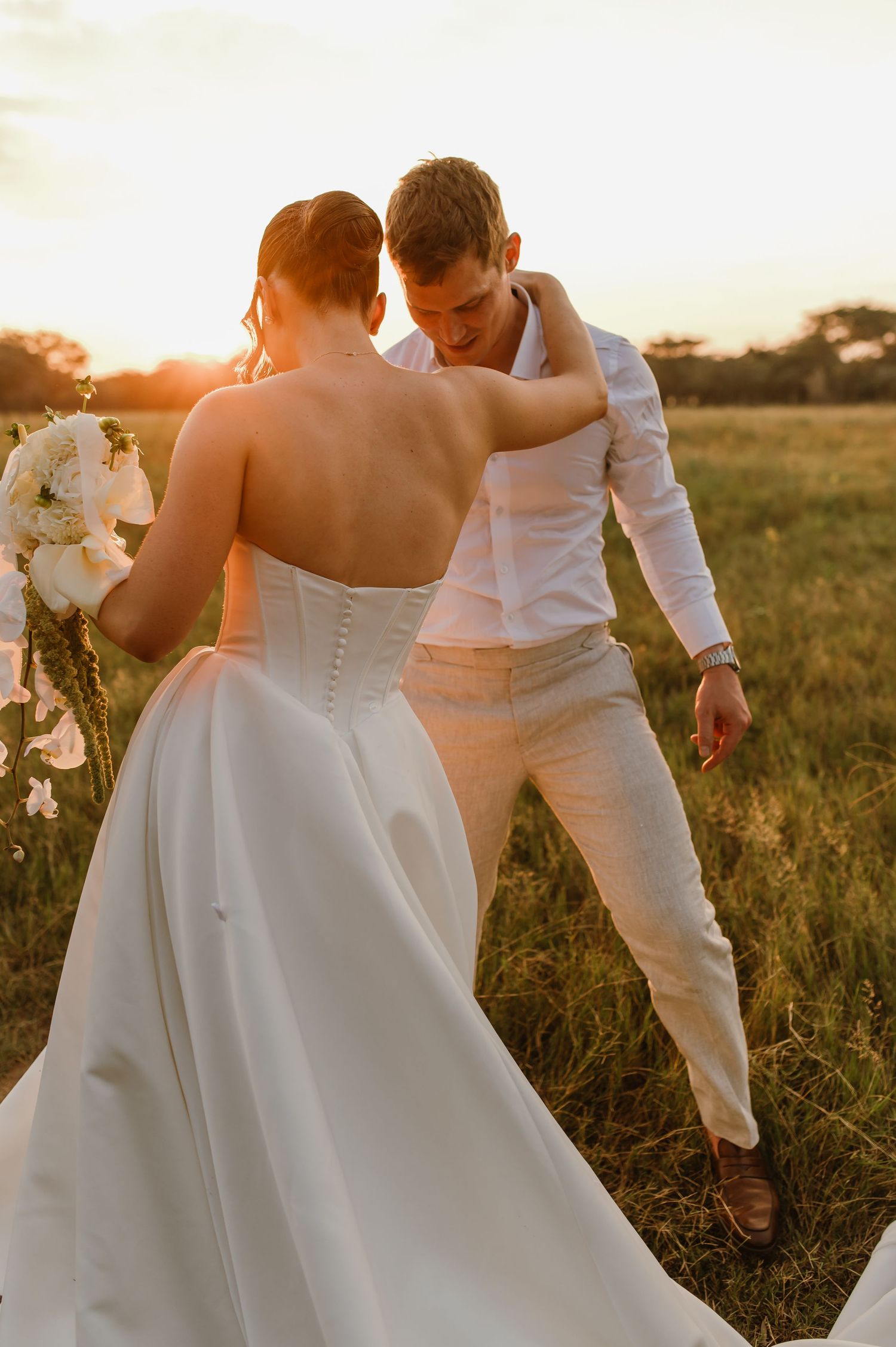 A couple walks hand in hand through tall grass during their sunset wedding shoot.