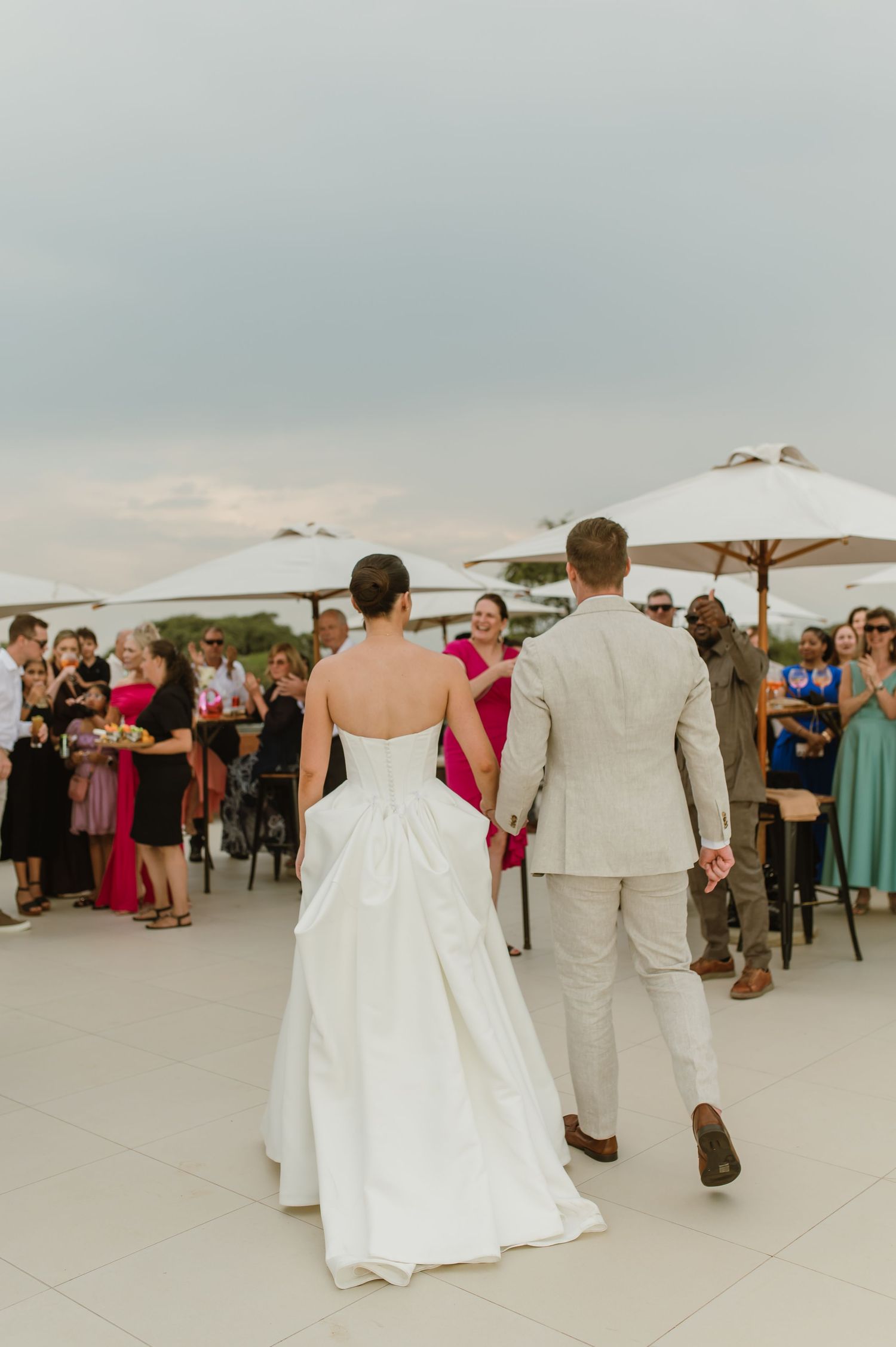 Wedding guests dance and celebrate on an outdoor patio with umbrellas and tables at sunset.