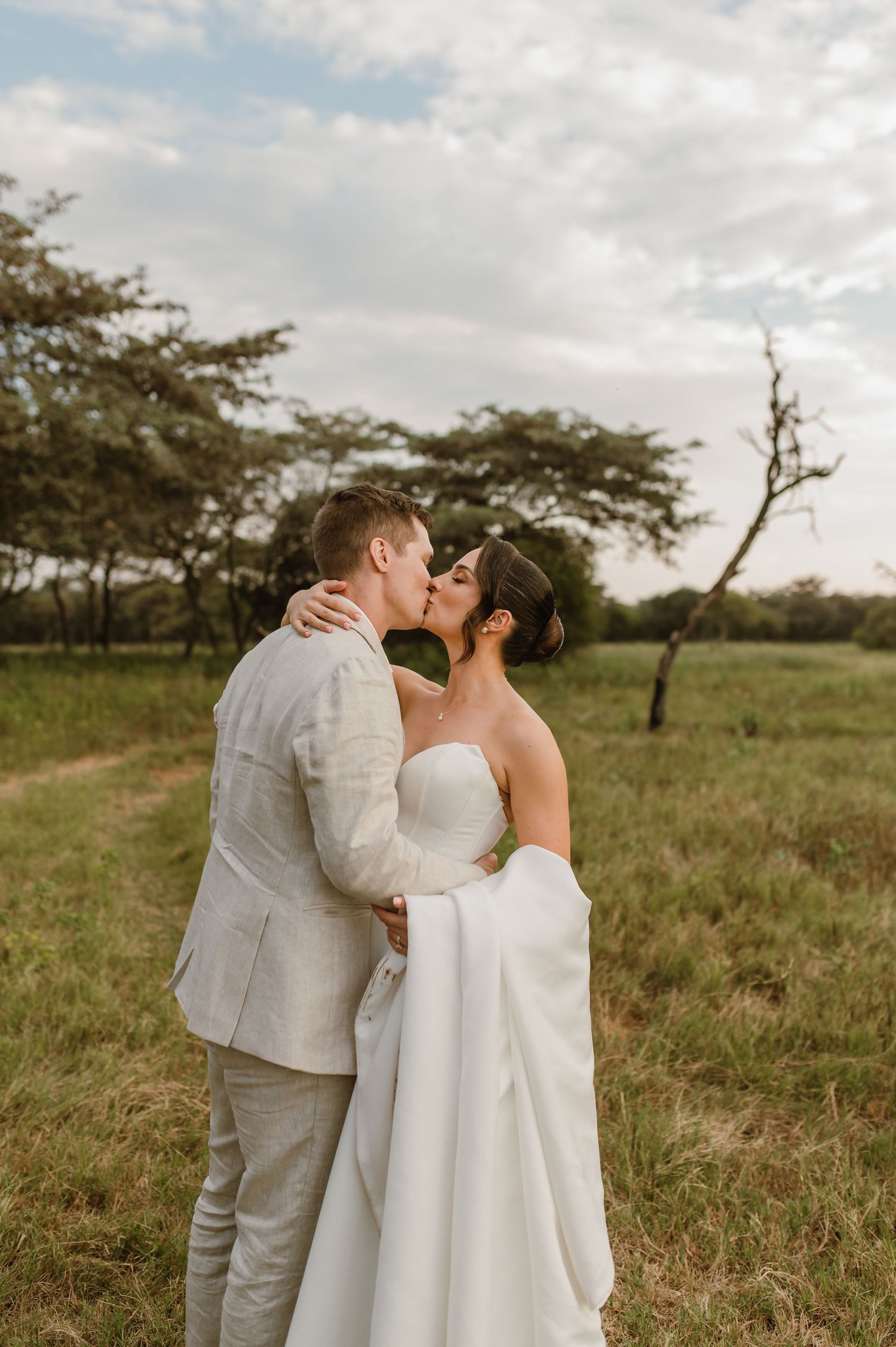 A couple shares an intimate moment in a grassy field with trees at sunset wearing white wedding attire.
