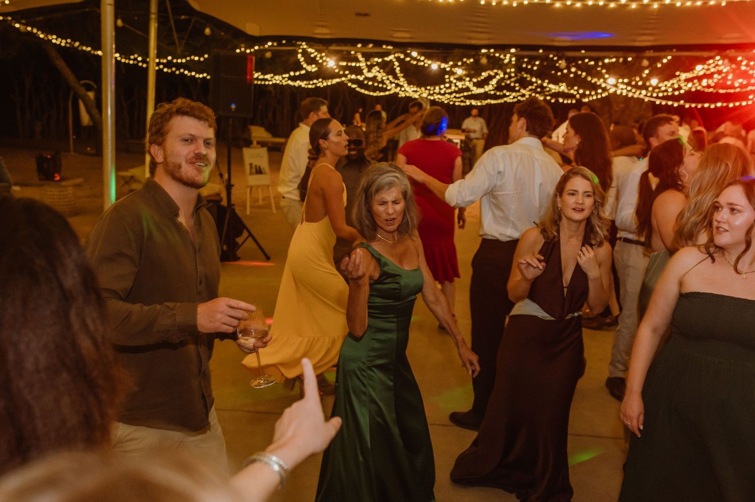 Wedding guests dancing and celebrating under warm string lights at an outdoor reception.