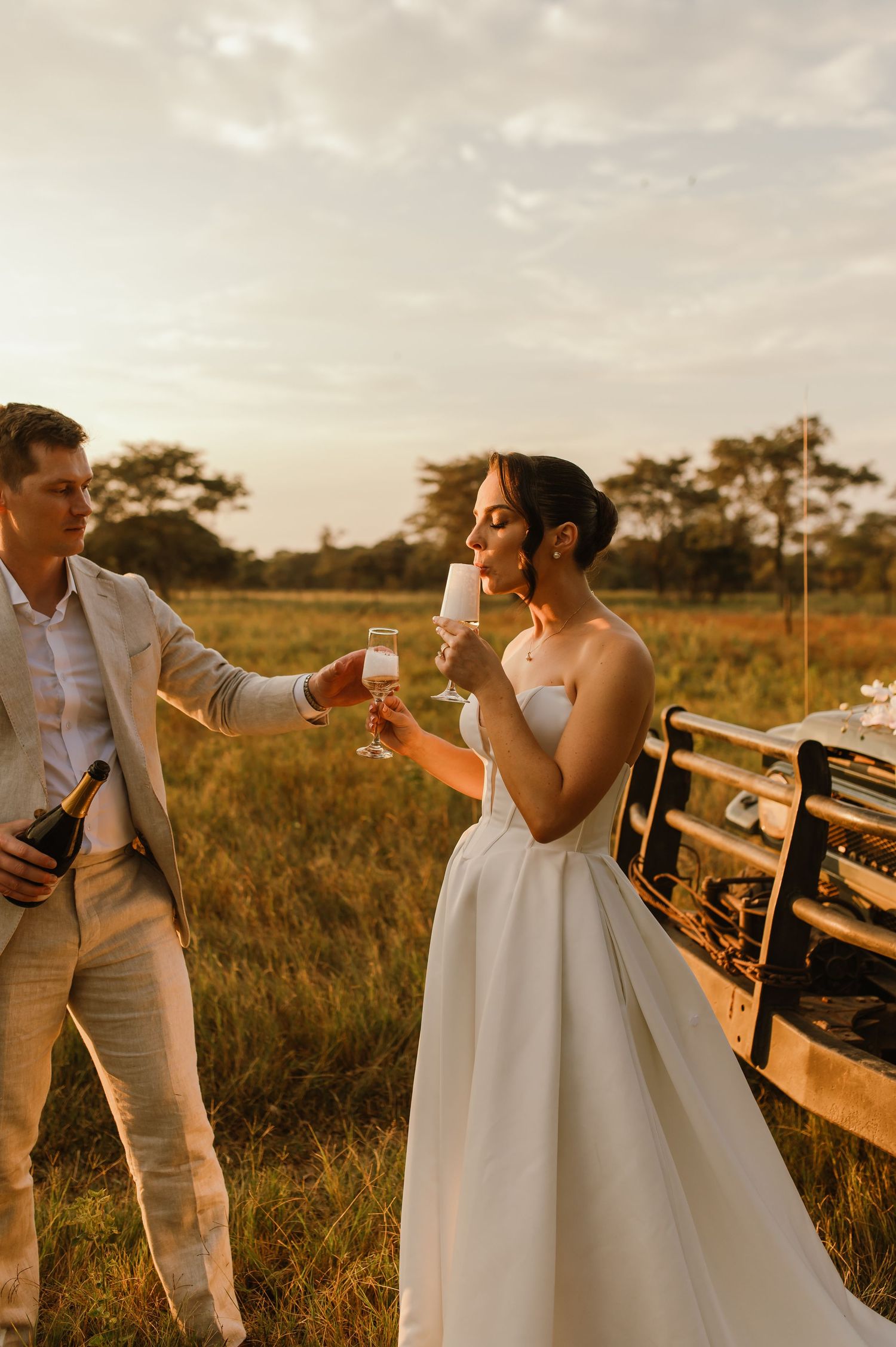 Playful wedding couple dancing and celebrating in a sun-lit field at sunset.