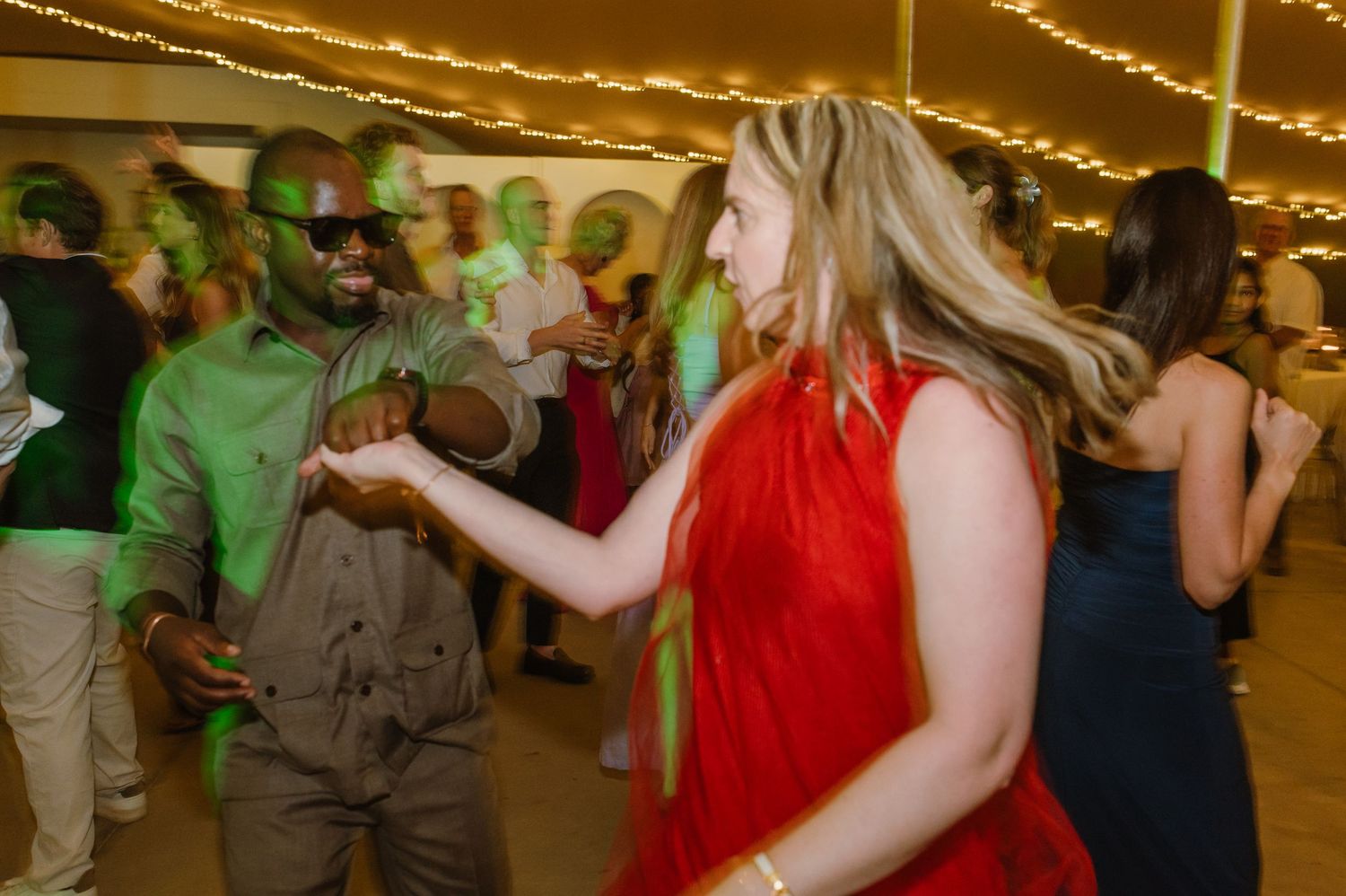 Wedding reception dance party with colorfully dressed guests celebrating under string lights.