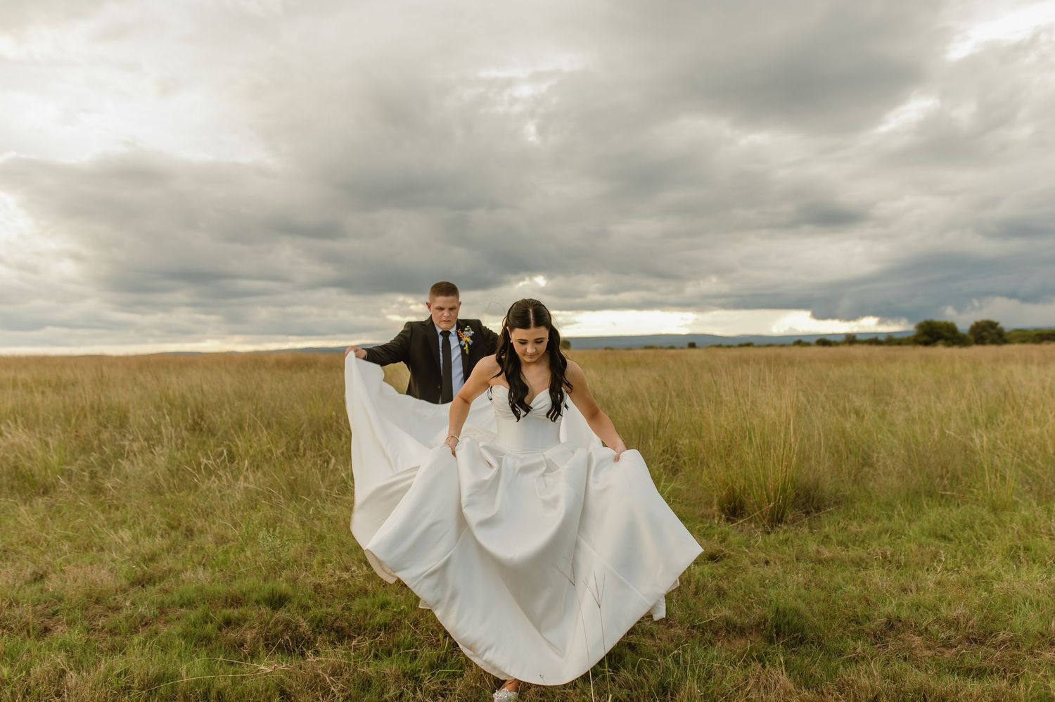 Bride and groom laughing together in an open field under dramatic cloudy skies.