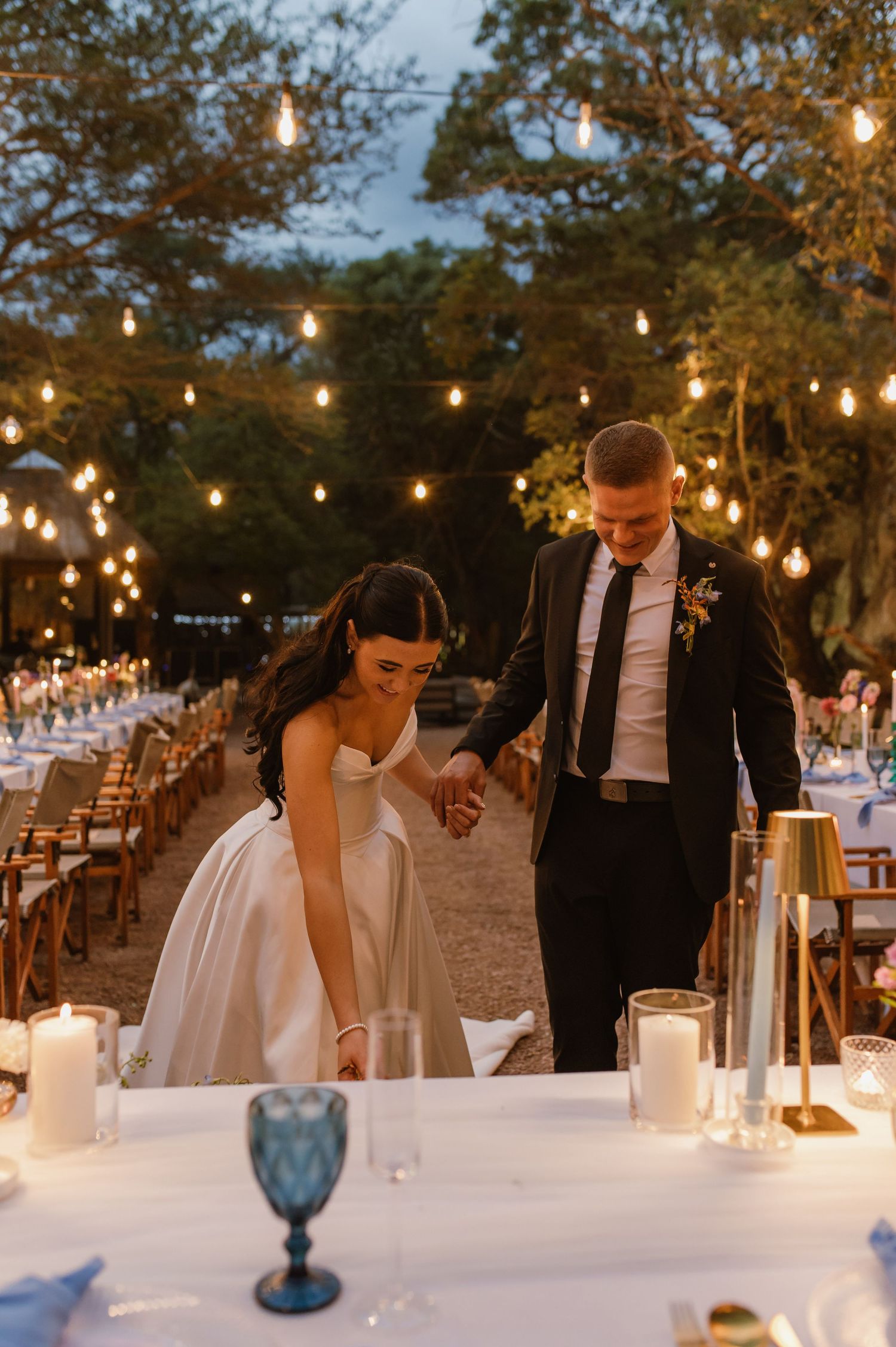 Bride and groom walk hand in hand at an elegant outdoor reception lit by warm string lights.