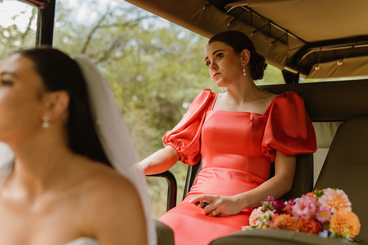 Bridesmaid in coral puff-sleeve dress holding floral bouquet seated in a vintage wedding transport.