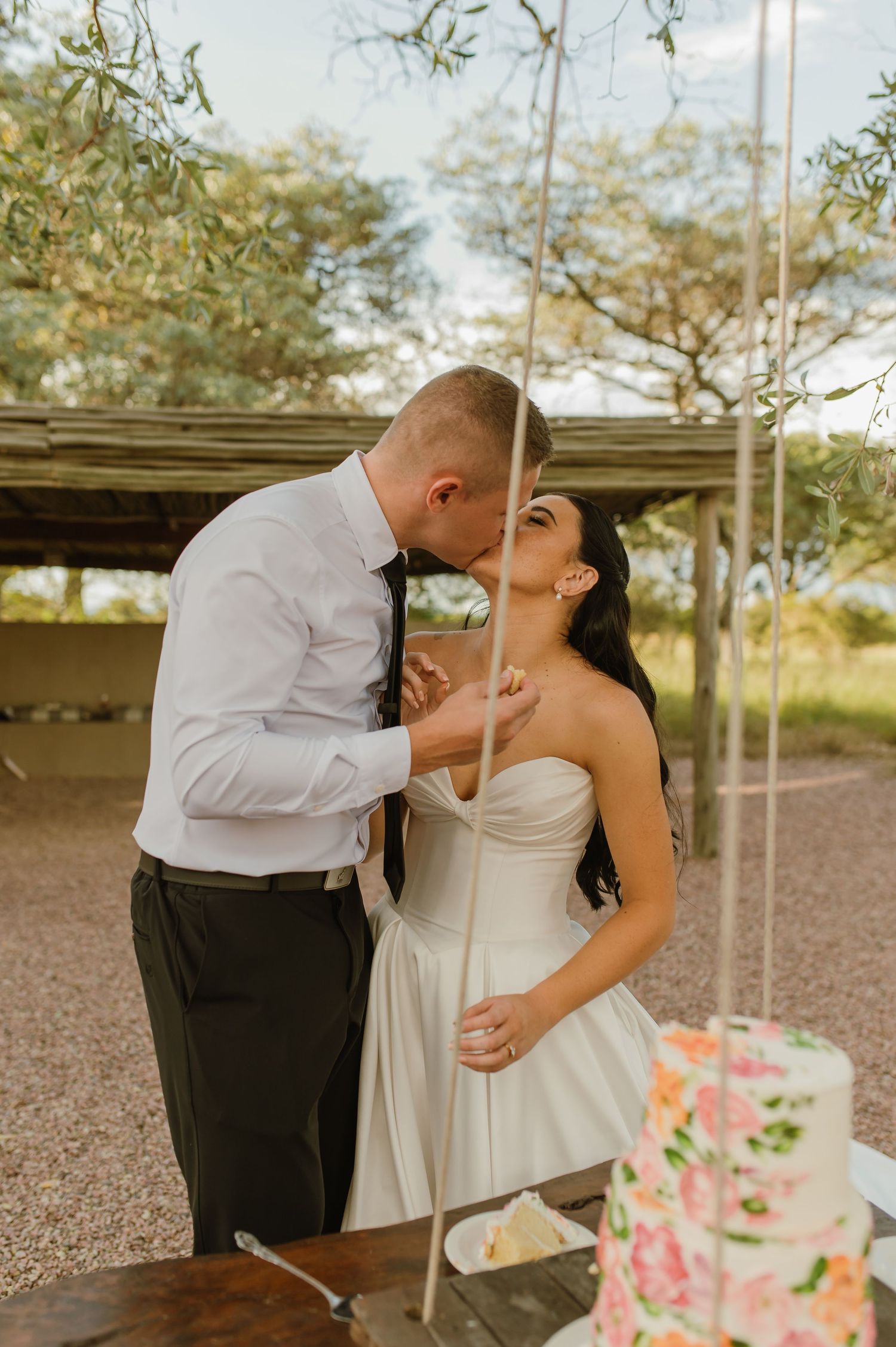 Bride and groom share a kiss while cutting their floral decorated wedding cake outdoors.