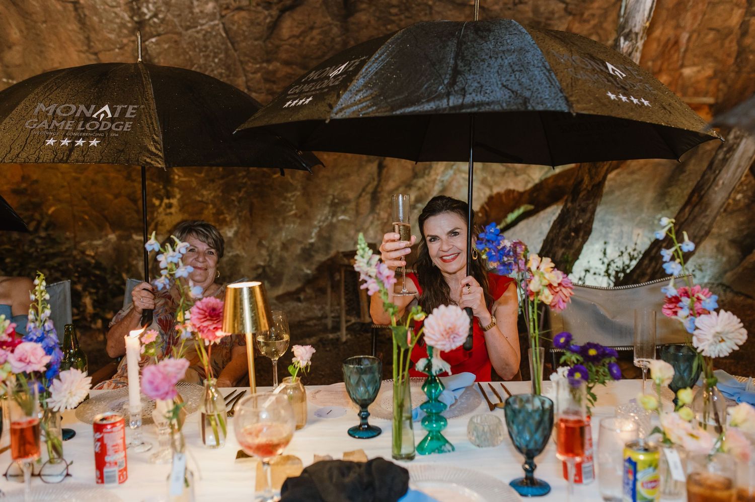 Woman in red dress smiling under black umbrellas at a colorful floral wedding reception table in a cave setting.