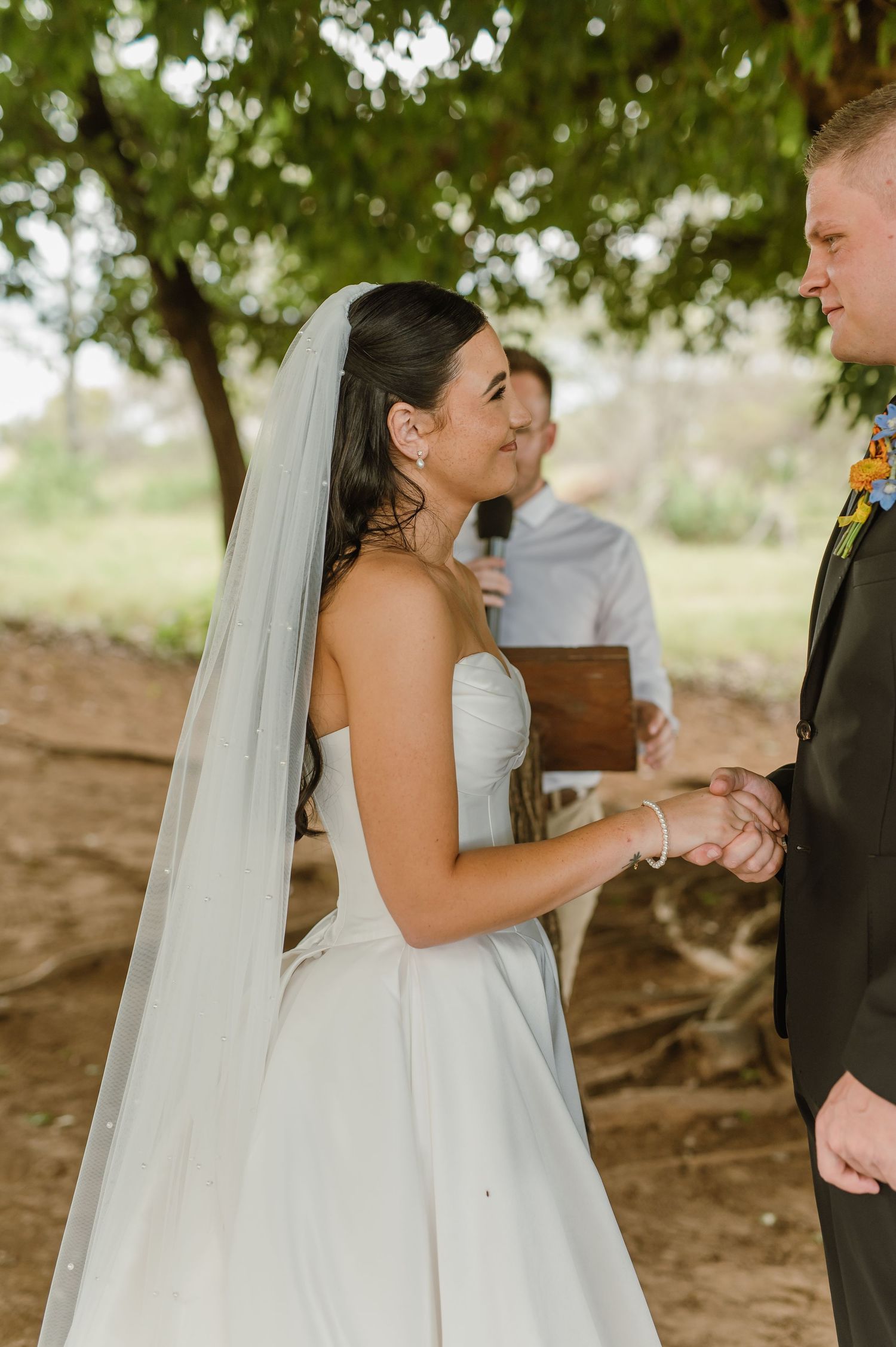 Bride in strapless white gown and veil holds grooms hands during outdoor wedding ceremony beneath a tree.