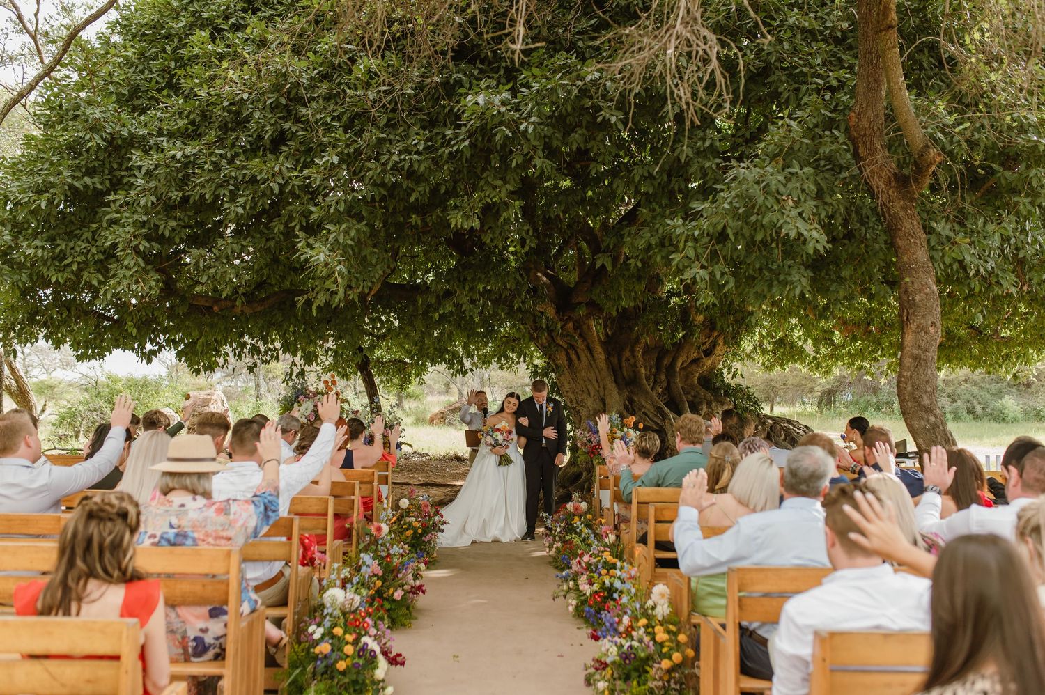 Outdoor wedding ceremony under a large tree with guests seated on wooden benches lining a floral aisle.