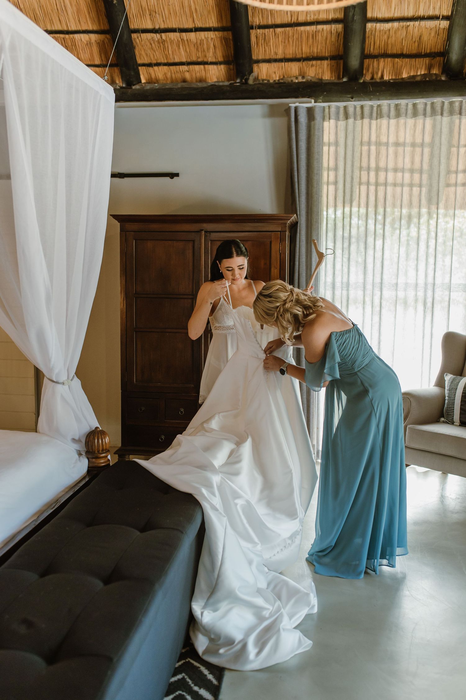 Bridesmaid in blue dress helping bride with white wedding gown in rustic thatched roof bedroom.
