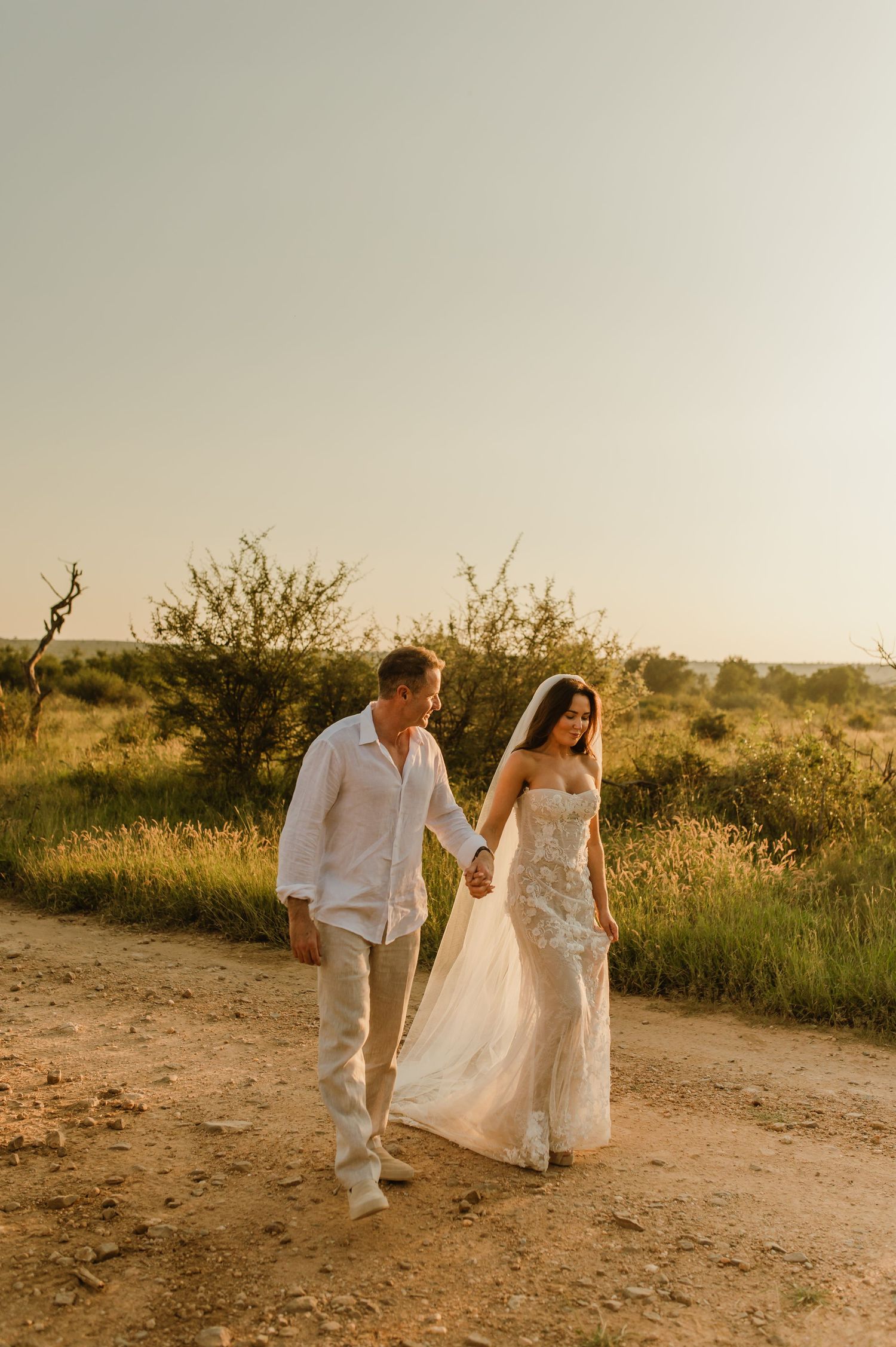 Bride in lace wedding dress and veil walks hand-in-hand with groom along sandy bush path at golden sunset.