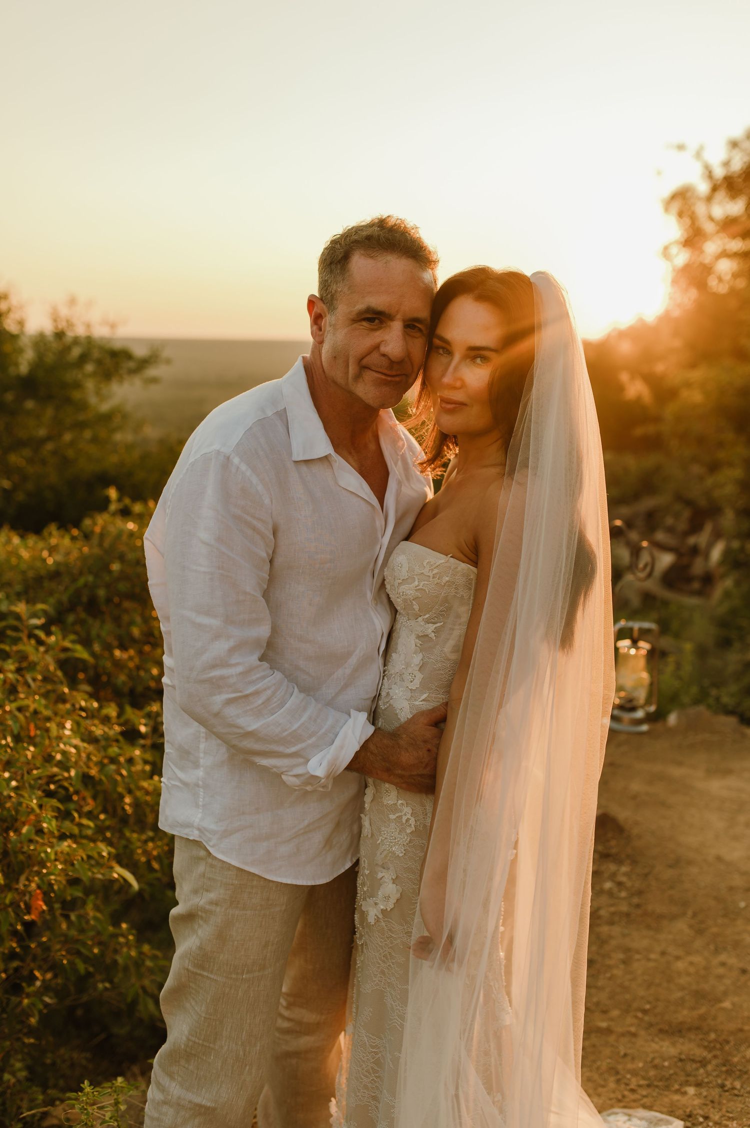 Bride in lace gown and veil embraces groom in white linen shirt during golden sunset outdoor wedding.