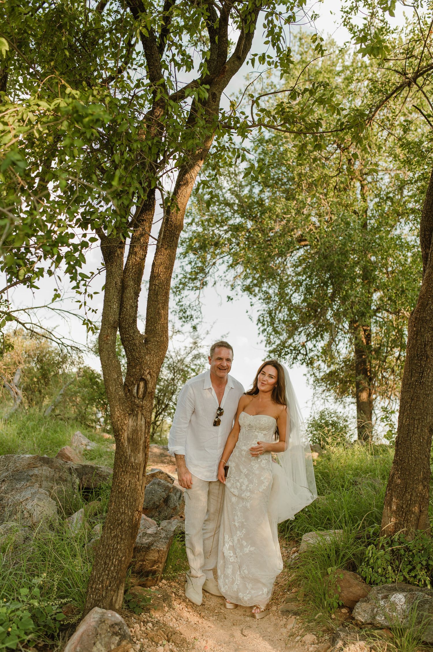 Bride and groom walking hand in hand through a lush tree-lined path on their wedding day.