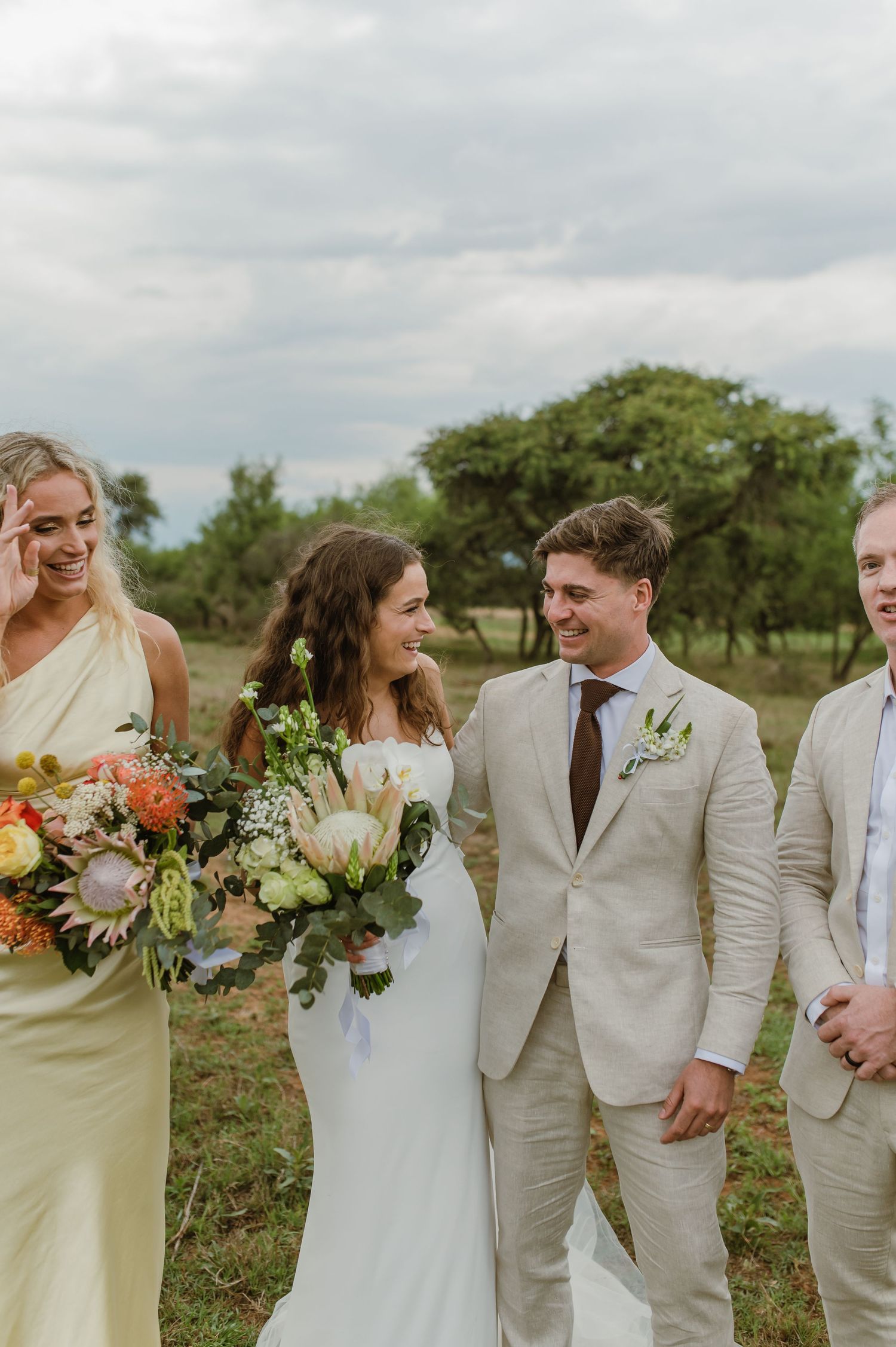 Wedding party in light colored attire posing together in rustic outdoor setting.