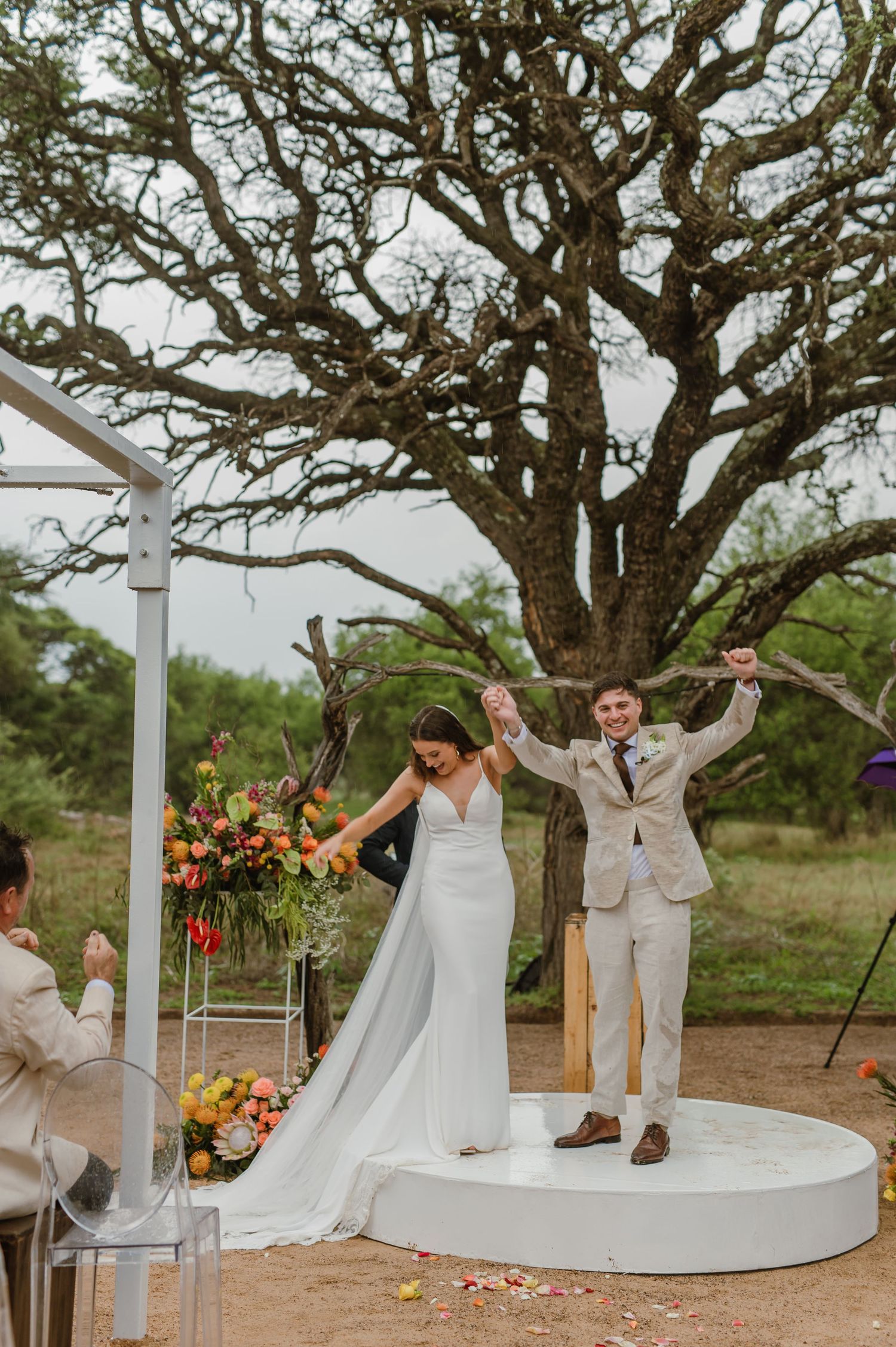 A couple celebrates with dynamic dance moves on a white circular platform during their wedding ceremony.