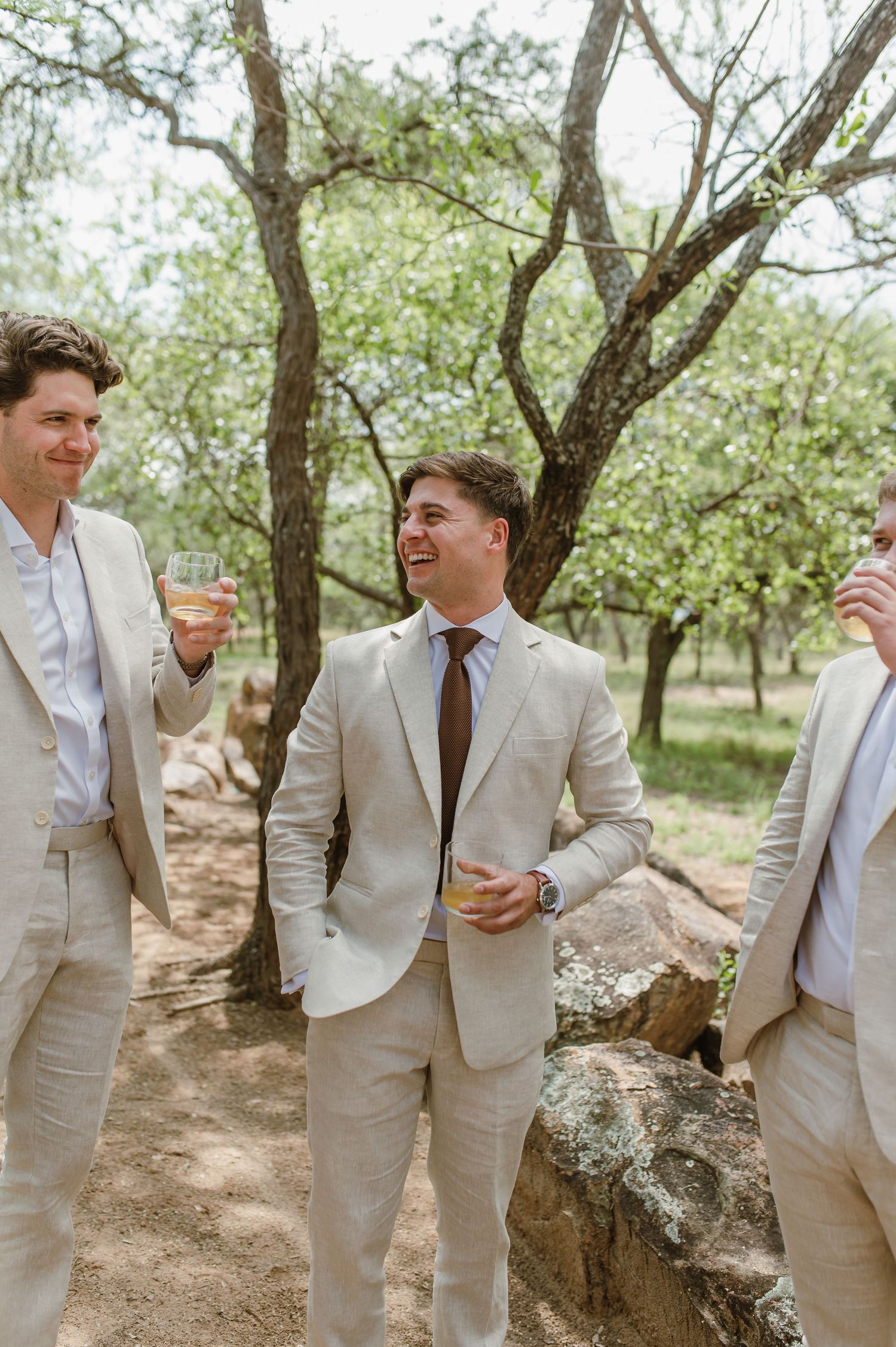 Wedding party members in beige suits share laughs among trees in a natural setting.