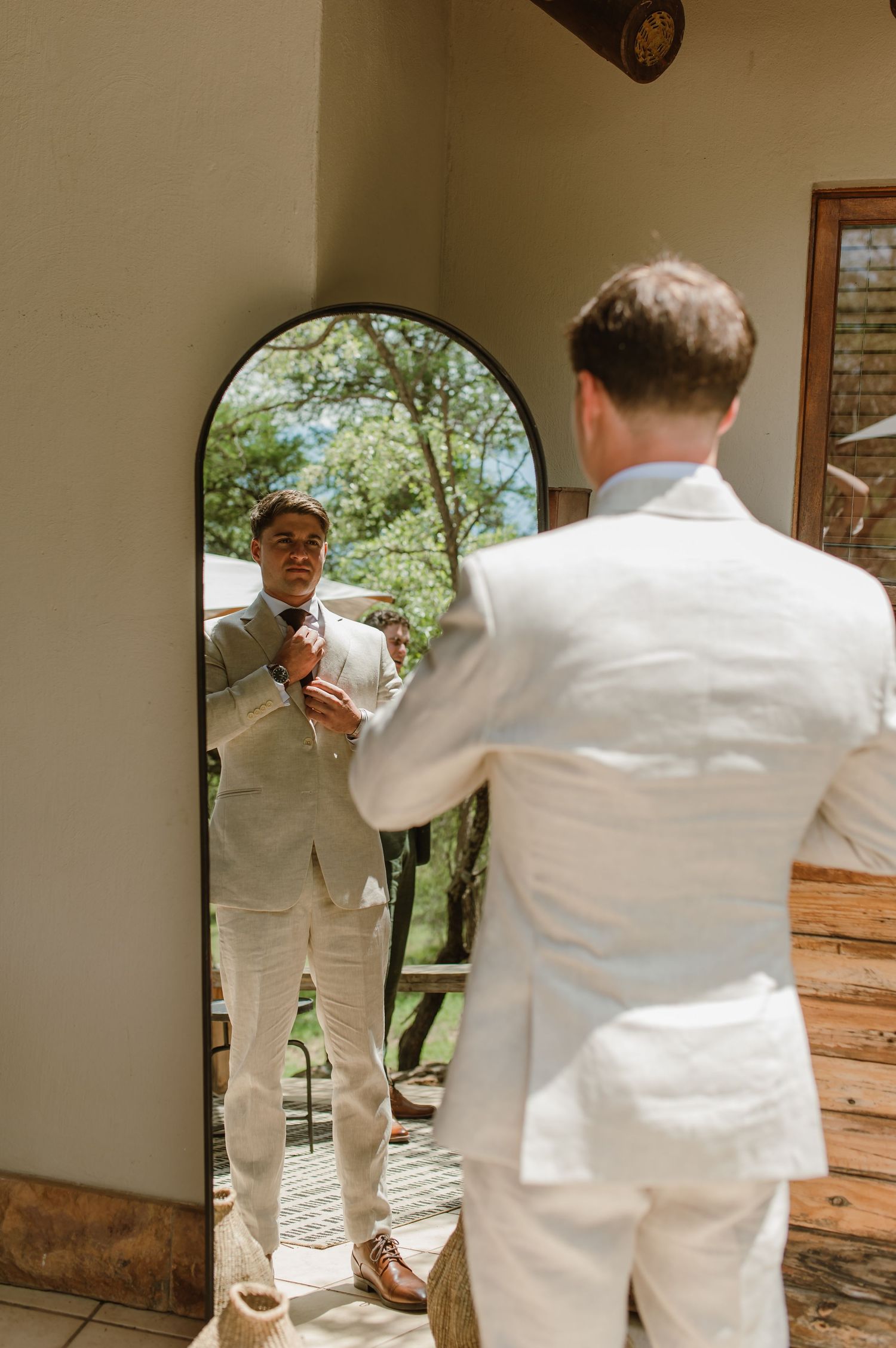 Man in white suit checks his appearance in a mirror during wedding preparations.