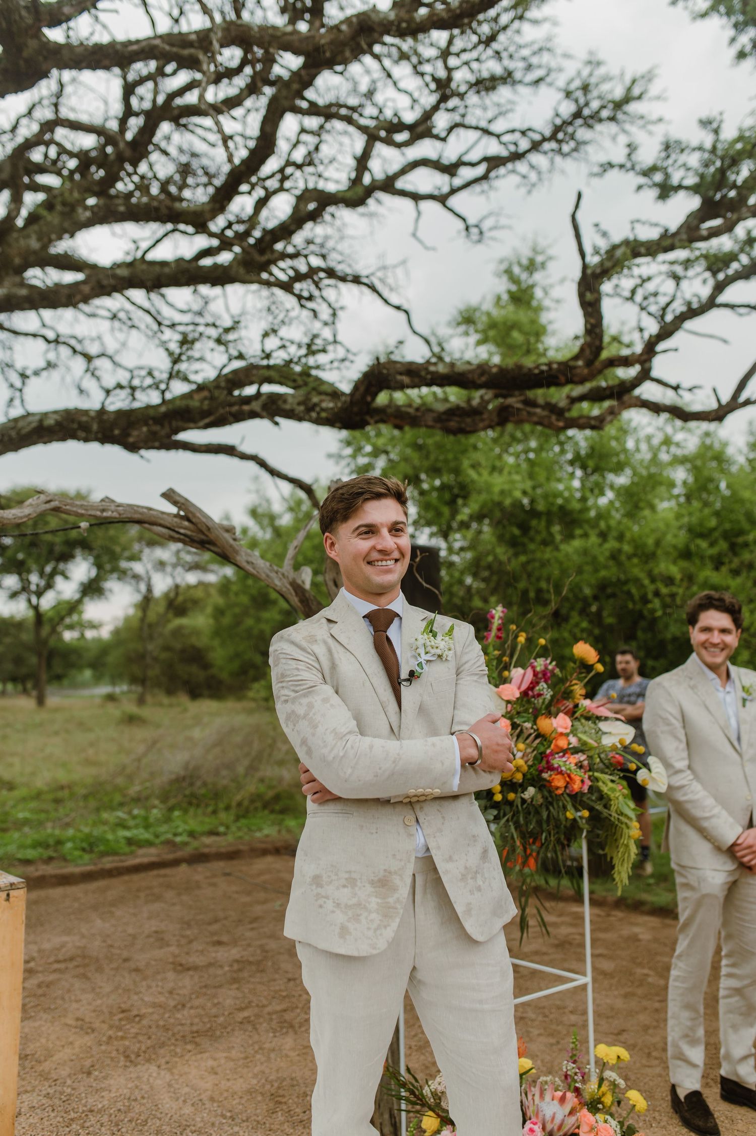 Groomsmen in light beige suits stand together under a large tree at an outdoor wedding ceremony.