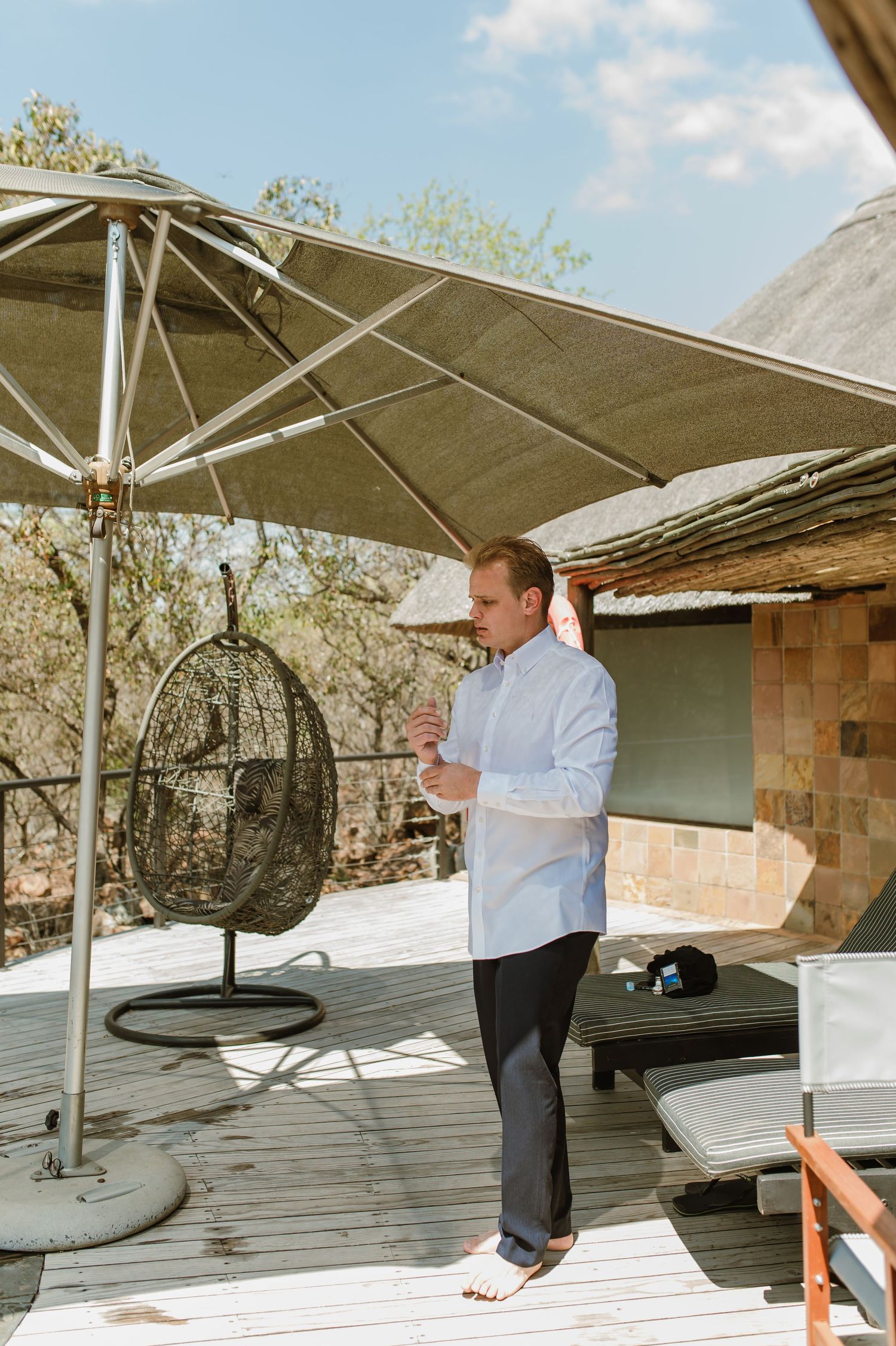 Someone in a white shirt stands on a deck with an umbrella and hanging chair at an outdoor resort setting.