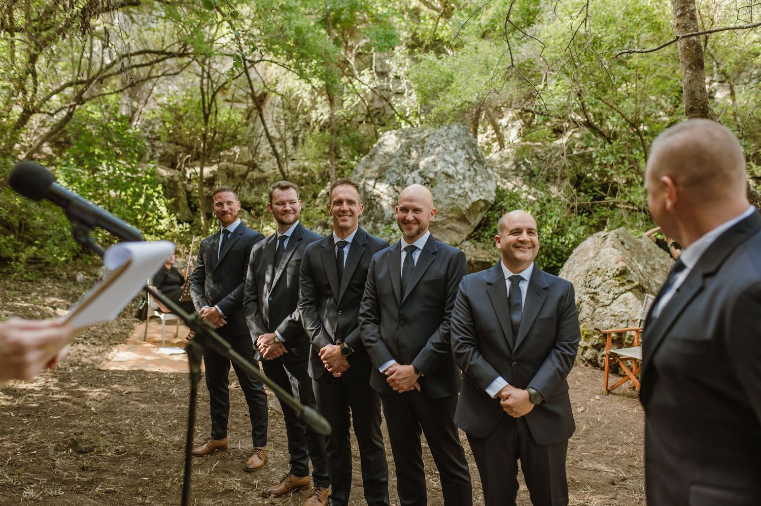 Groomsmen in dark suits gather for wedding ceremony photos in wooded setting.