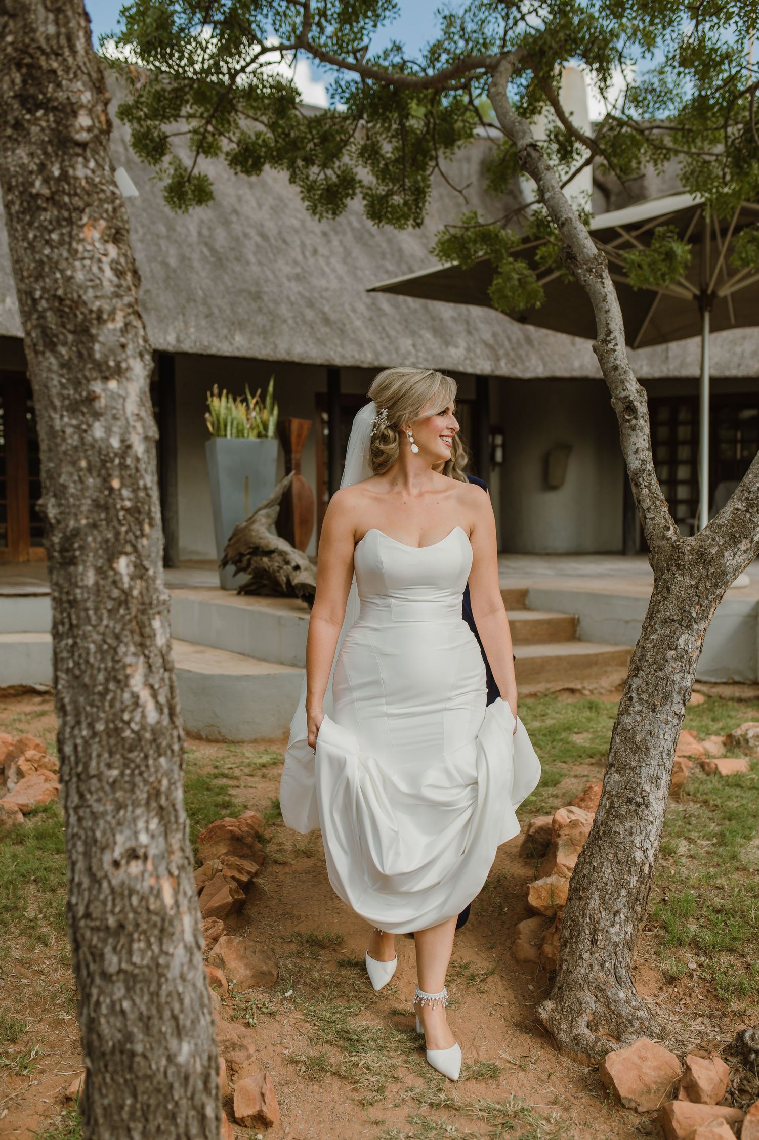 A bride in a white wedding dress poses playfully between trees in an outdoor garden setting.