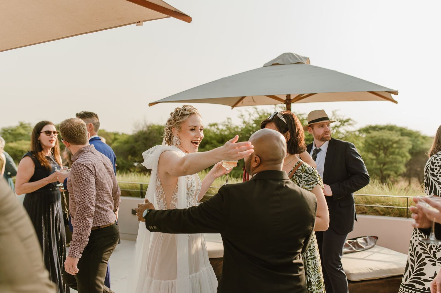 Outdoor wedding reception with guests mingling under beige umbrellas at sunset.