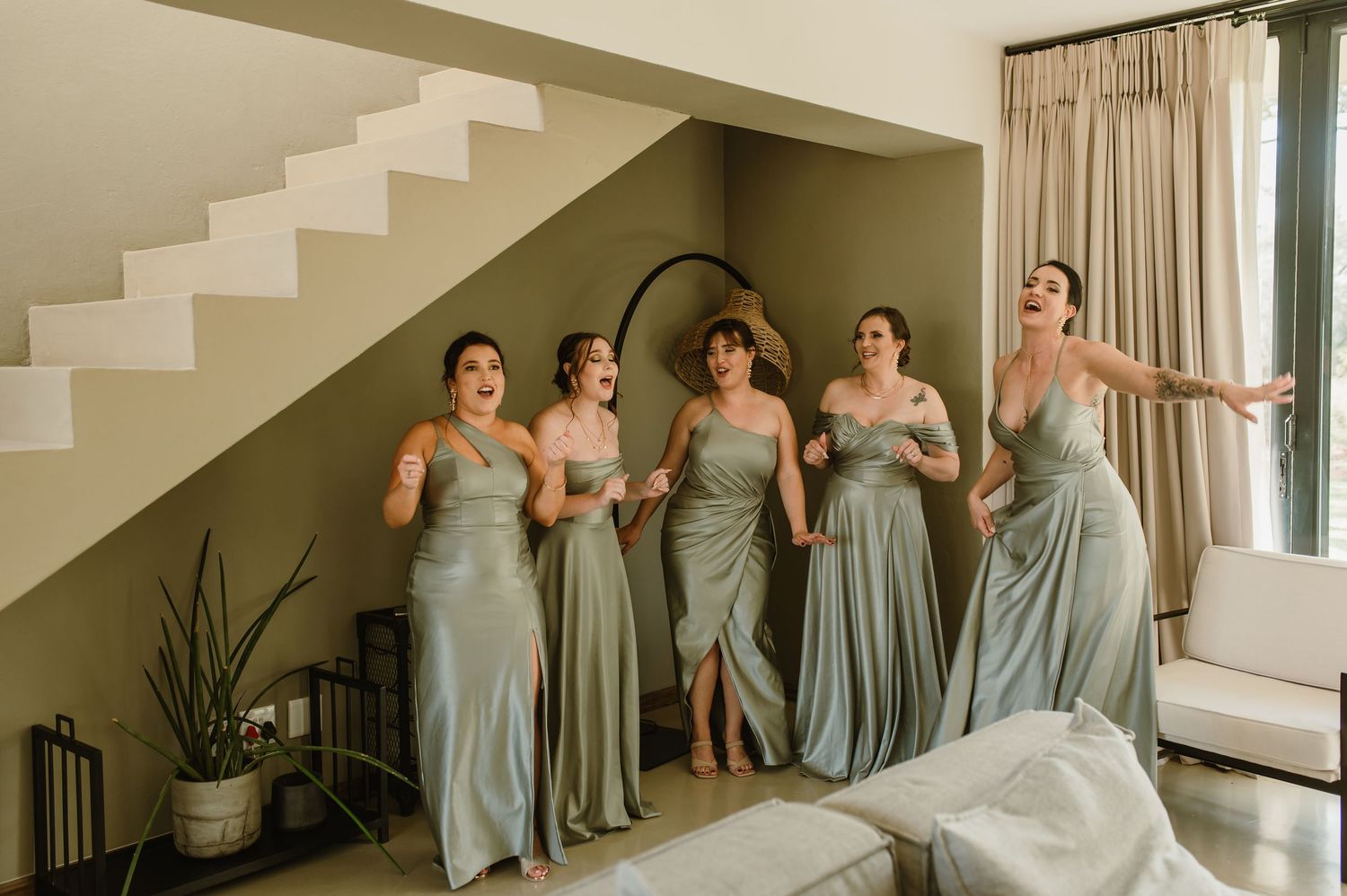 Bridesmaids in sage green dresses react emotionally on a modern staircase.