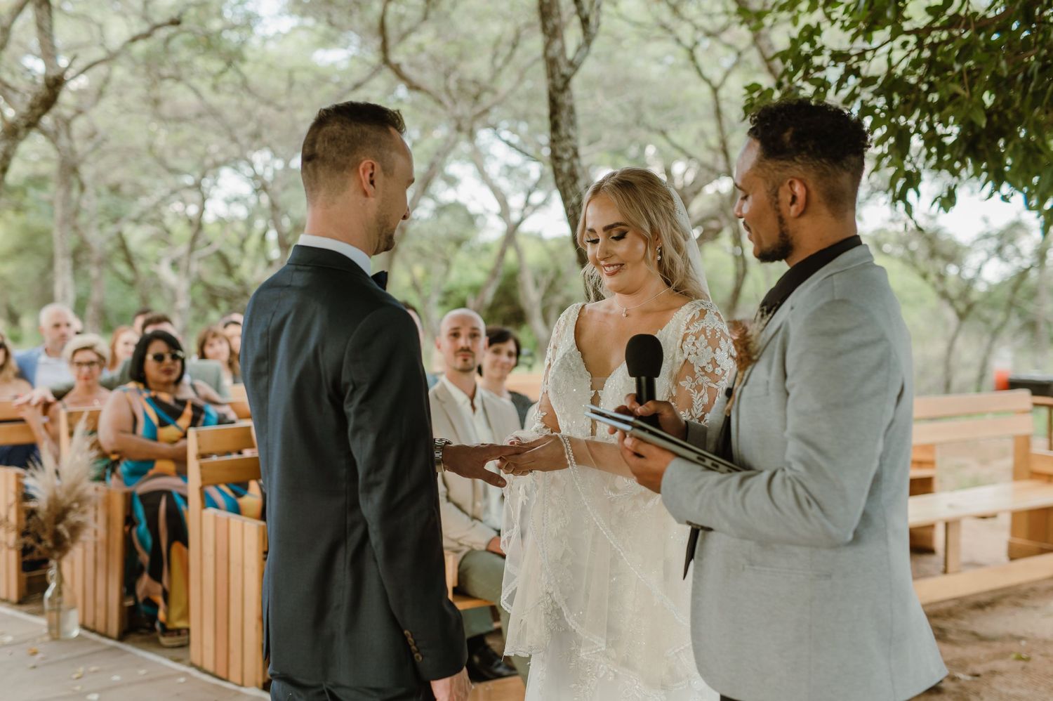 An outdoor wedding ceremony takes place with wooden bench seating under tree coverage.