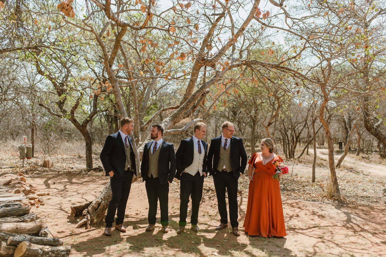 A wedding party in formal attire poses together under bare trees in a desert landscape.