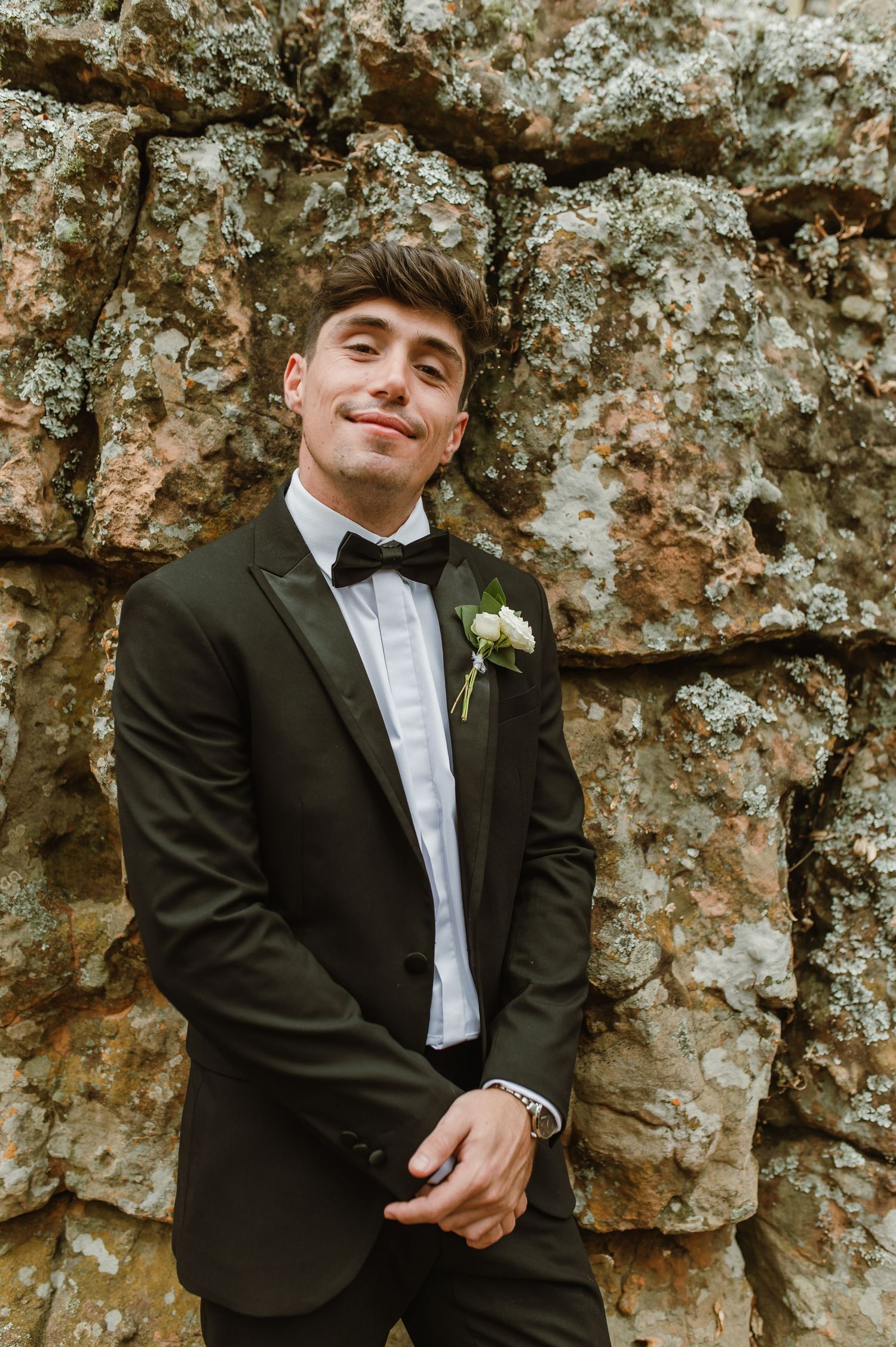 Groomsman in black tuxedo and bow tie poses against textured rock wall with boutonniere.