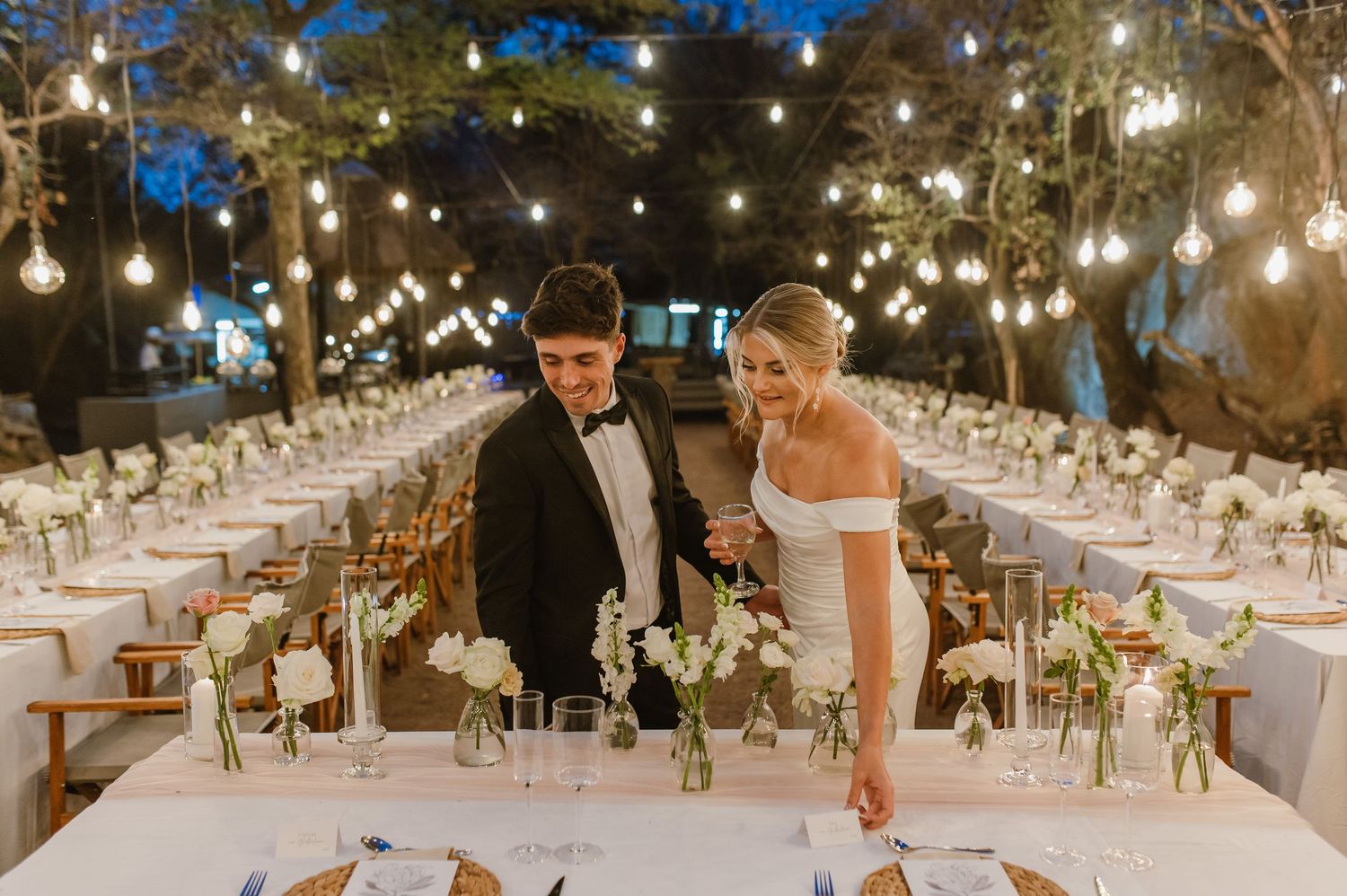 Elegant outdoor dining setup with rows of white-clothed tables under twinkling string lights.
