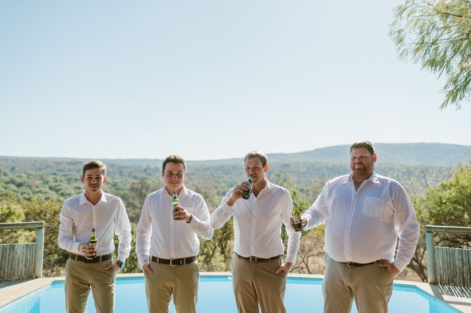 Four groomsmen in casual attire standing by pool with mountain backdrop.