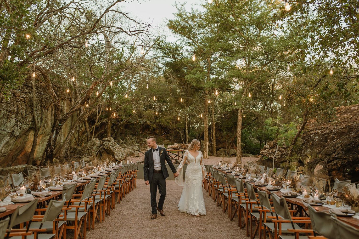 A couple walks down an outdoor reception aisle lined with wooden chairs and hanging lights.