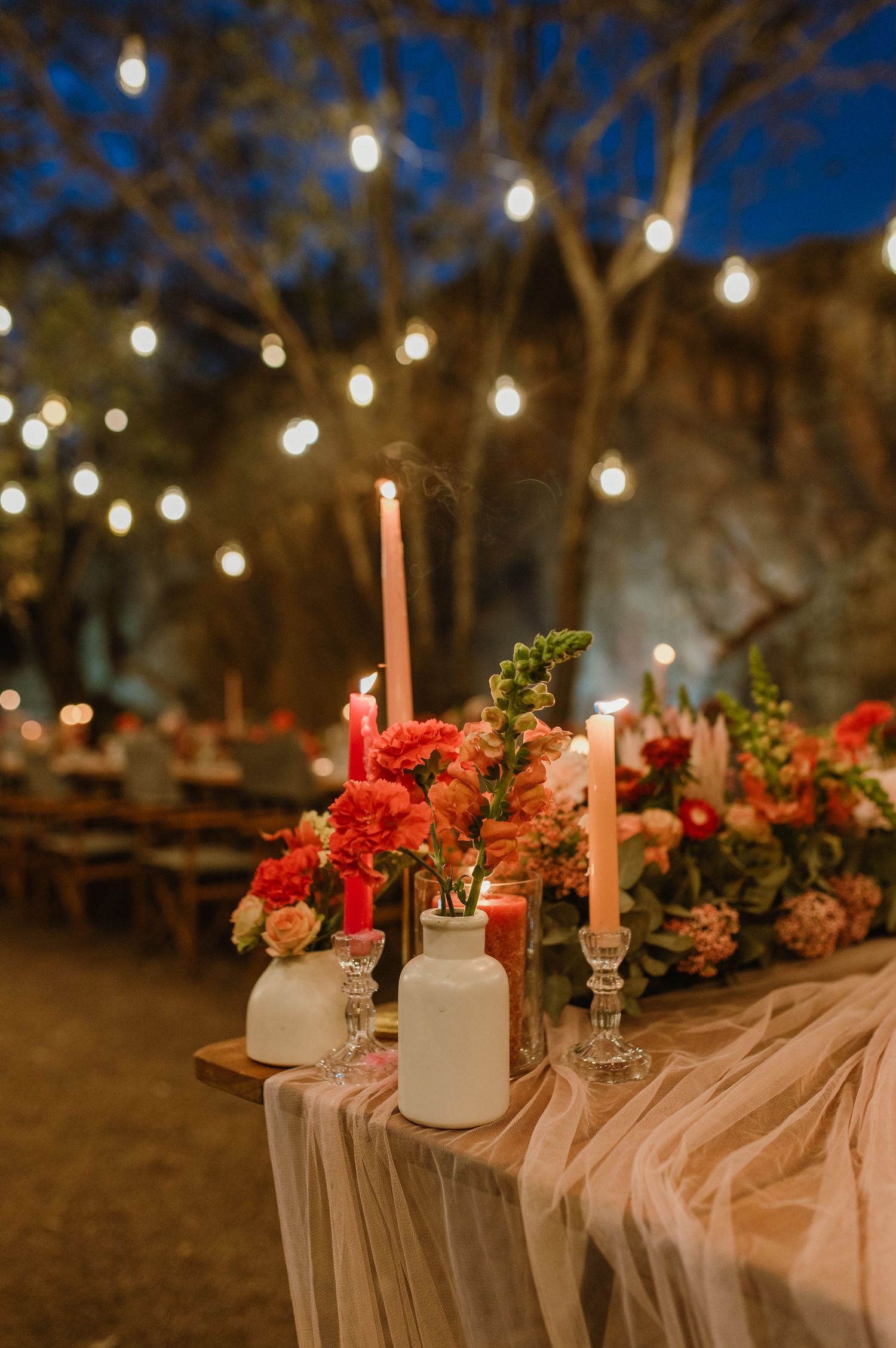 Wedding table decorated with pink draping, red flowers and tall candles under string lights.